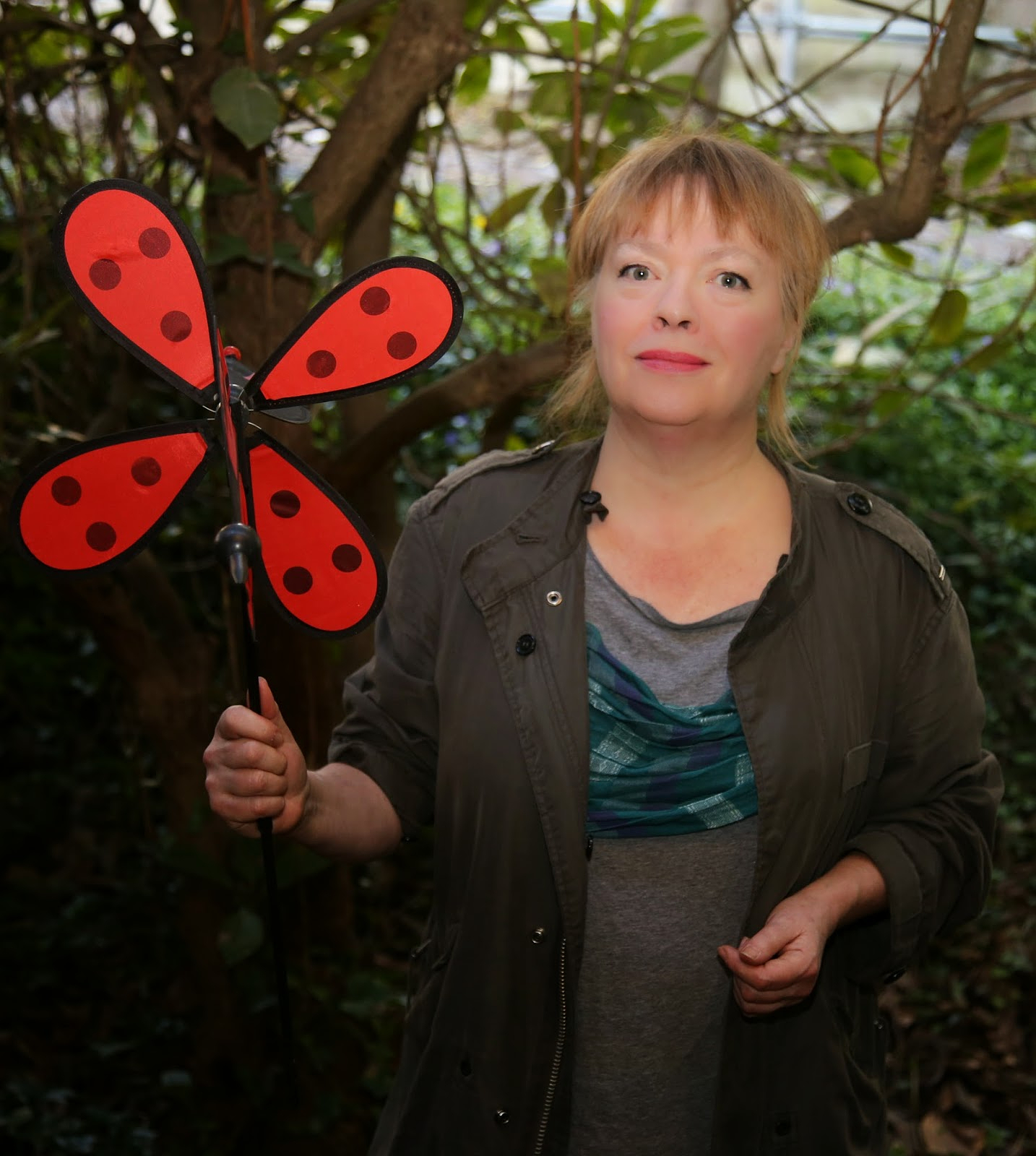 Smiling, Dorothy Trujillo Lusk stands in front of a tree and holds a ladybug-spotted pinwheel in her left hand. She wears an olive jacket open over a grey dress.