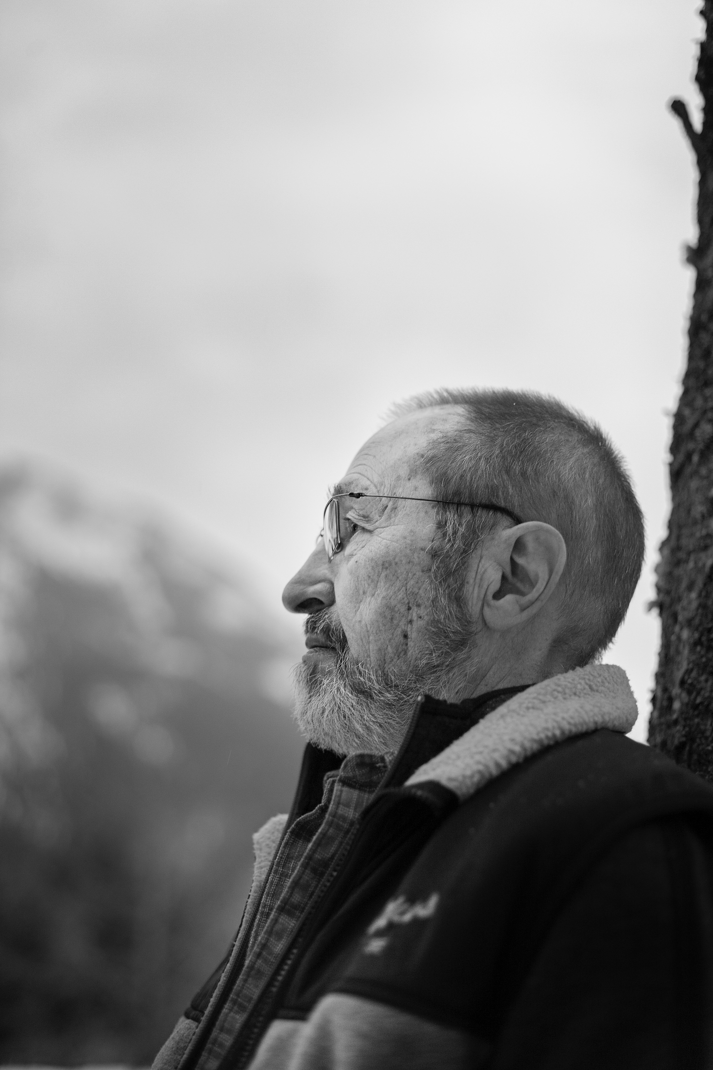 Seen in profile, Fred Wah wears glasses and a jacket with a sherpa collar in this black-and-white portrait. A cloudy mountain is visible in the background.