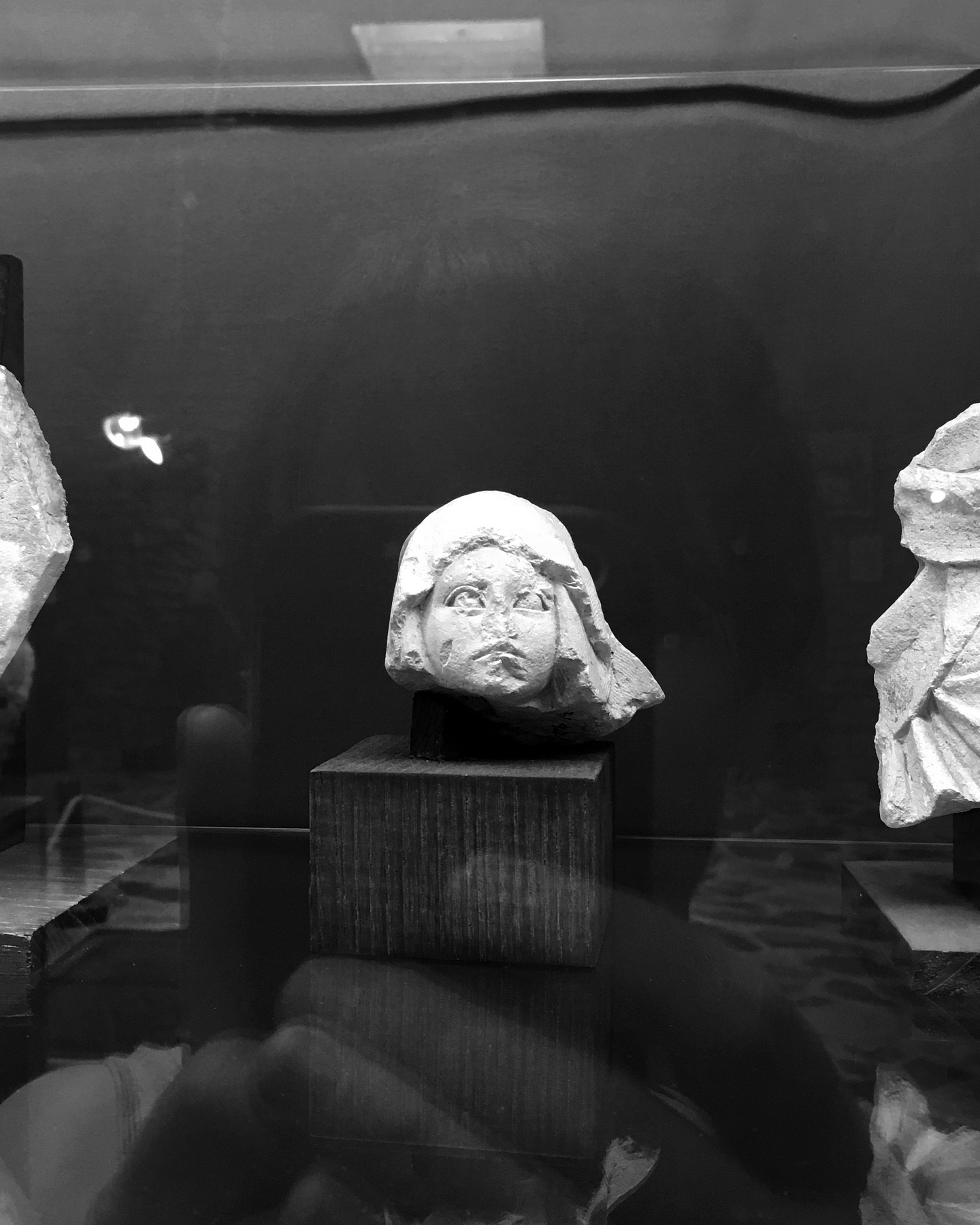 A black-and-white mirror selfie taken in the reflection of a museum display case holding a small sculpture of a head.