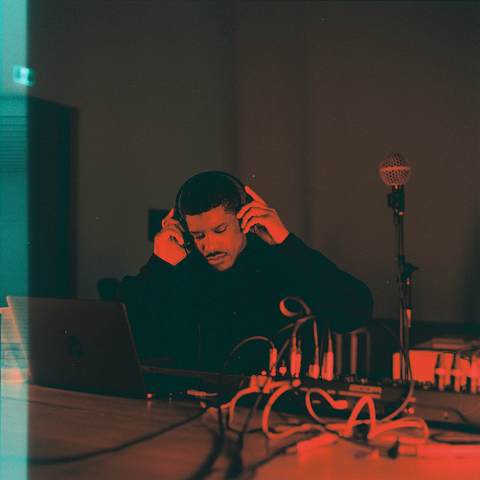 DeForrest Brown, Jr. works at a table of electronic music equipment in a dim, red-lit room, wearing headphones and surrounded by a laptop, mixer, coiled cables, and a microphone on a stand. The photograph, shot on film, reveals a visible exposure along the edge of the frame.