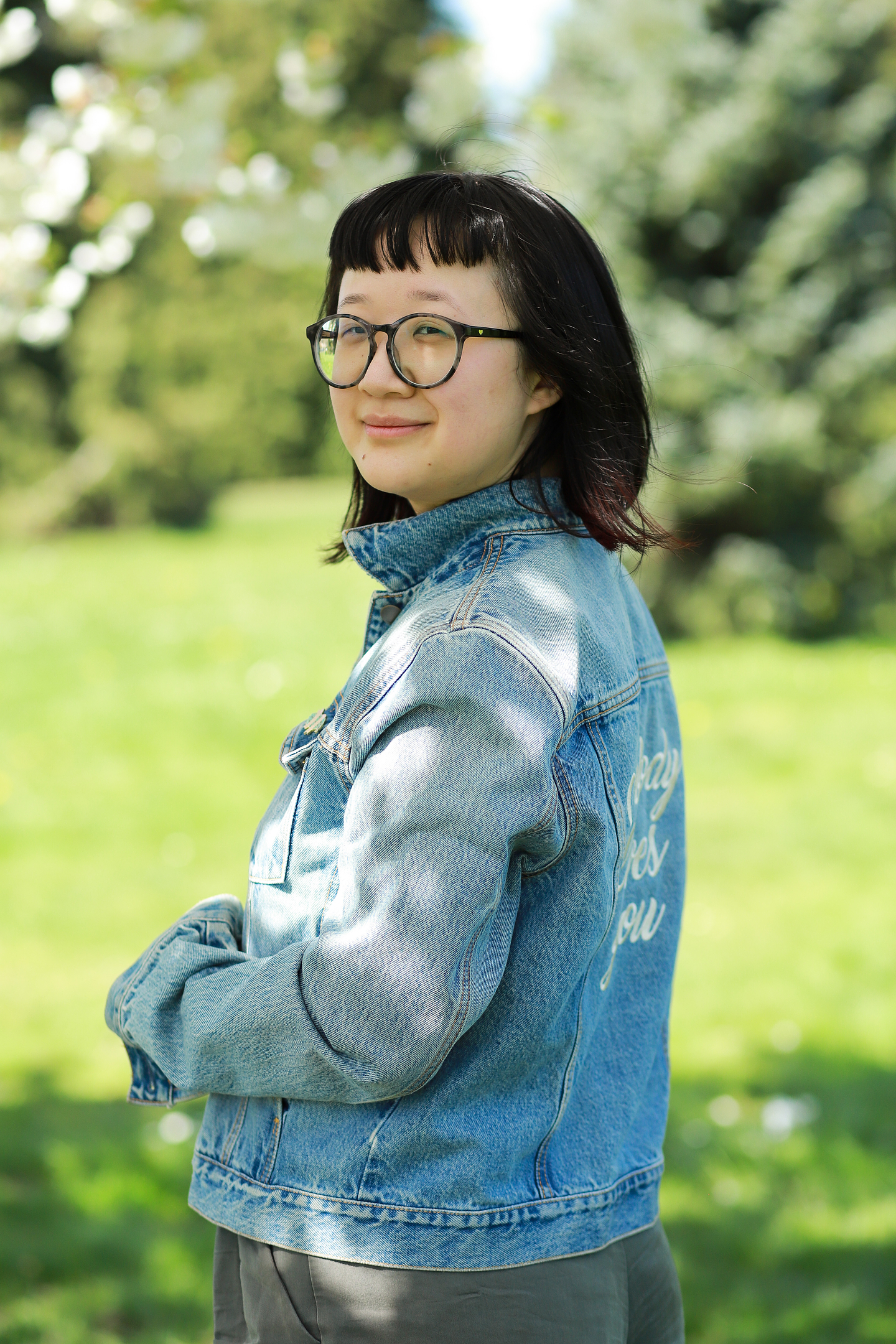 Jane Shi stands outside and looks over her shoulder to smile at the camera. She wears a denim jacket with text embroidered on the back. In the background lush grass and green foliage can be seen out of focus.
