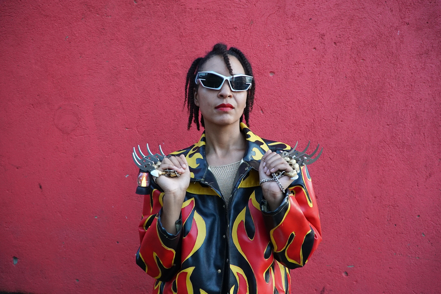 Portrait of Violence Gratuite standing against a red wall, wearing a black leather jacket with bold red and yellow flame patterns. She has short twisted hair, angular sunglasses, red lipstick, and hold ups spiked metal accessories in both hands, exuding a confident and defiant pose.