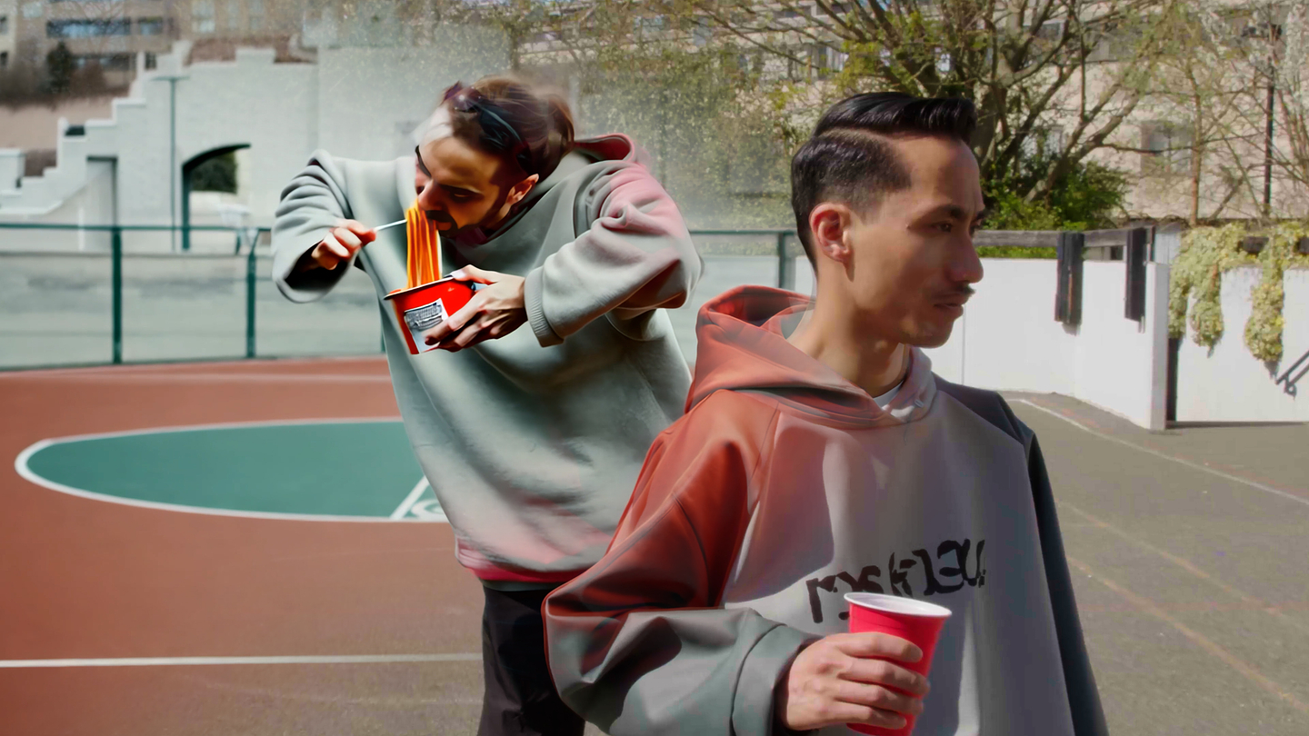 The image shows two people on an outdoor basketball court; one in the background is hunched over enthusiastically eating noodles from a red cup, while the other in the foreground faces away, casually holding a red cup and wearing a hoodie with text on it. The scene appears bright and candid, with both individuals dressed in oversized sweatshirts and the court and surrounding walls visible behind them.