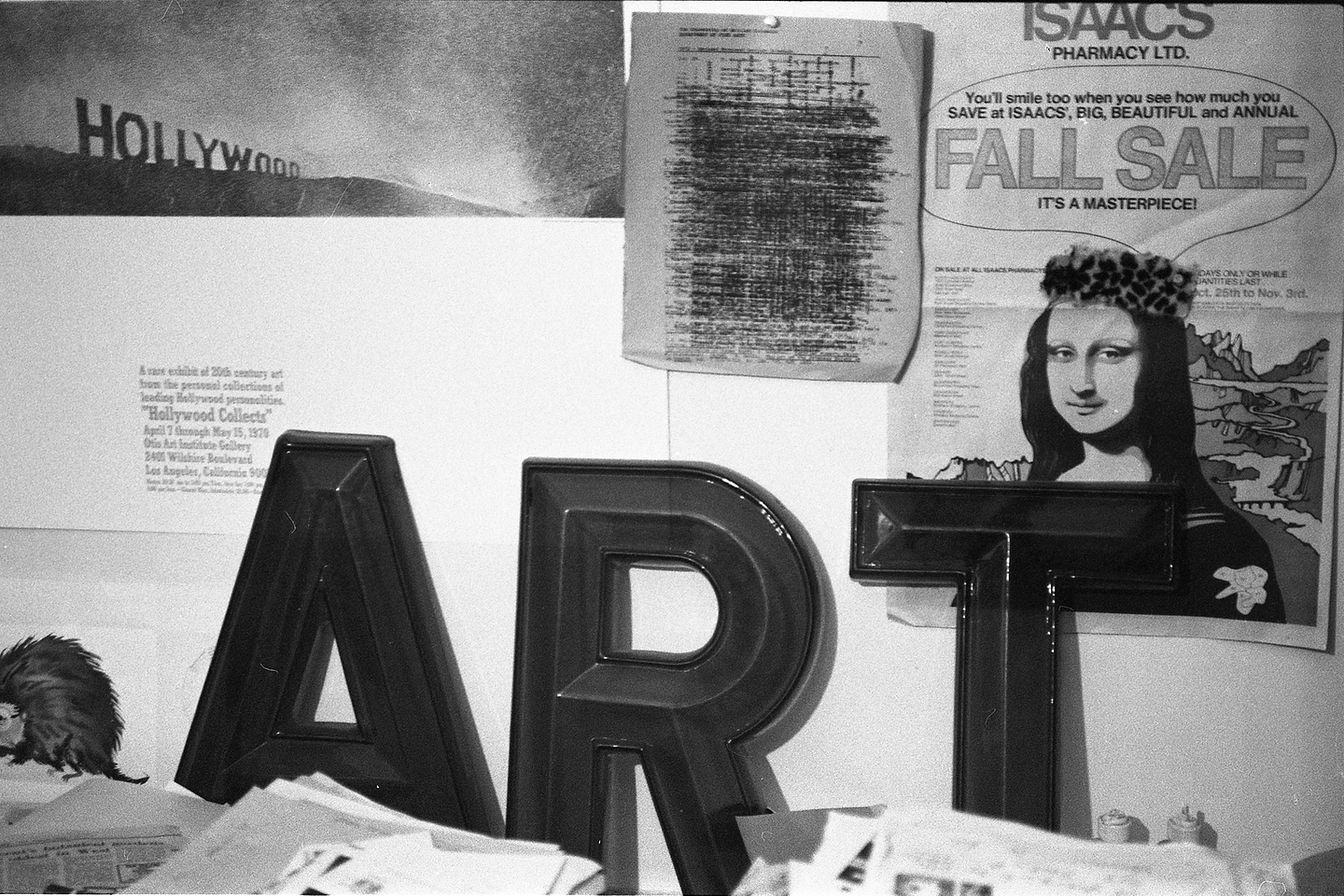 A black and white photograph of large sculptural black letters spelling out “A R T” leaning against a wall. The wall has posters and other pieces of paper tacked up on it, including an advertisement for Isaacs Pharmacy Ltd that features an image of Da Vinci’s Mona Lisa with text that reads: “You’ll smile too when you see how much you SAVE at ISAAC’S, BIG, BEAUTIFUL, and ANNUAL FALL SALE: IT’S A MASTERPIECE!”, and an exhibition poster for “Hollywood Collects” that shows a photograph of the Hollywood sign at sunset. More stacks of paper are visible in the foreground.