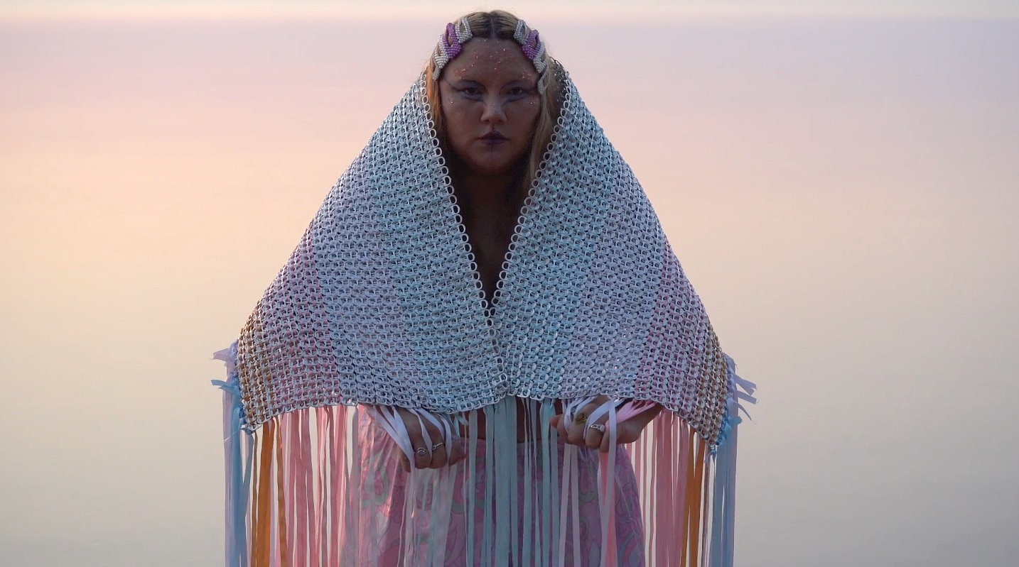 Cheyenne Rain LeGrande looks directly at the camera, while wearing a  fringed shawl made by weaving pastel coloured ribbons through aluminum can tabs. Her face is adorned with face paint and her hair is pinned back with decorated clips. She stands outside and is illuminated by the soft glow of the setting sun.