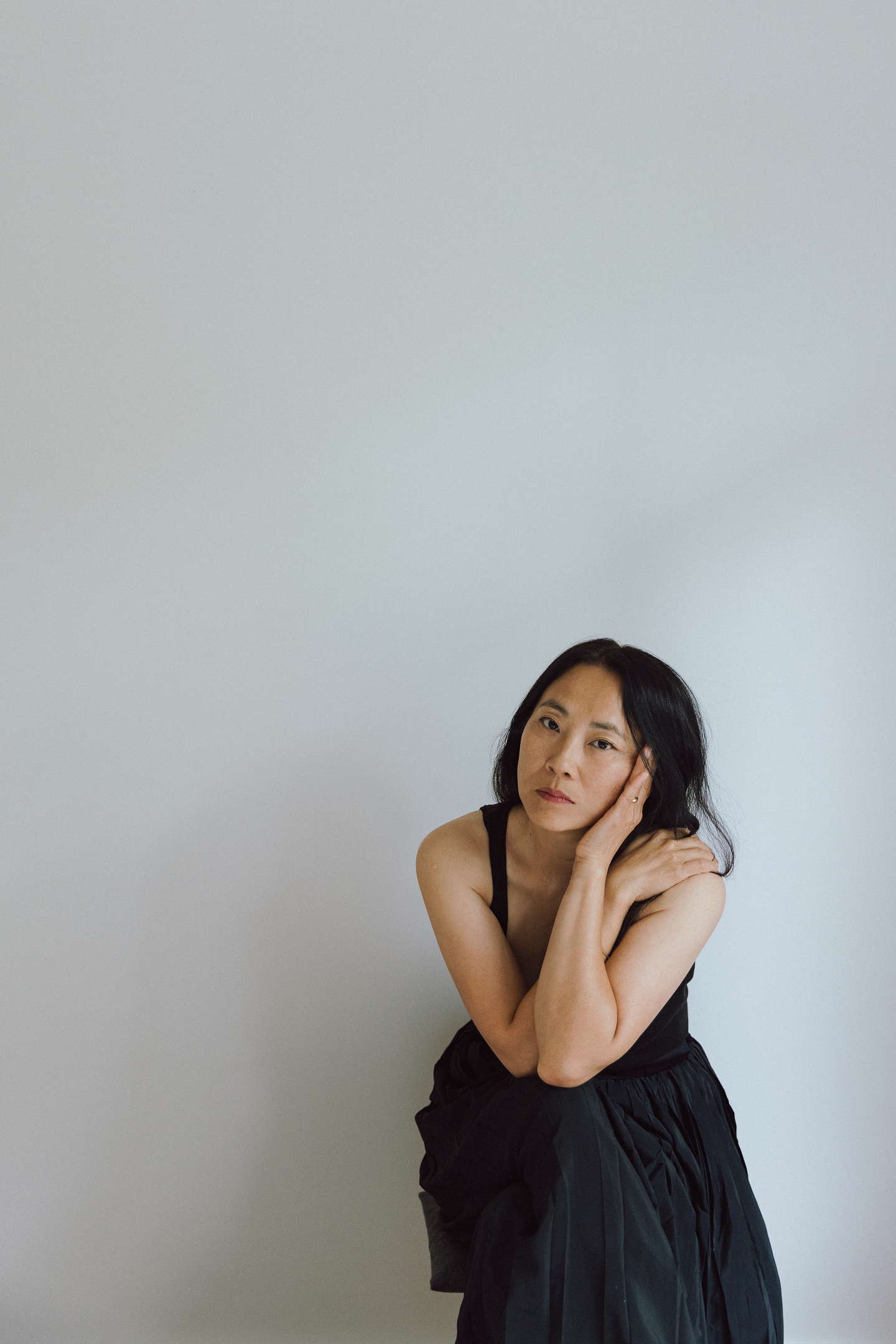 Yoon Sook Cha wears a black sleeveless dress and sits in front of a white wall. She leans forward, resting her elbows on the knee of her crossed leg while her hand cradles her head.