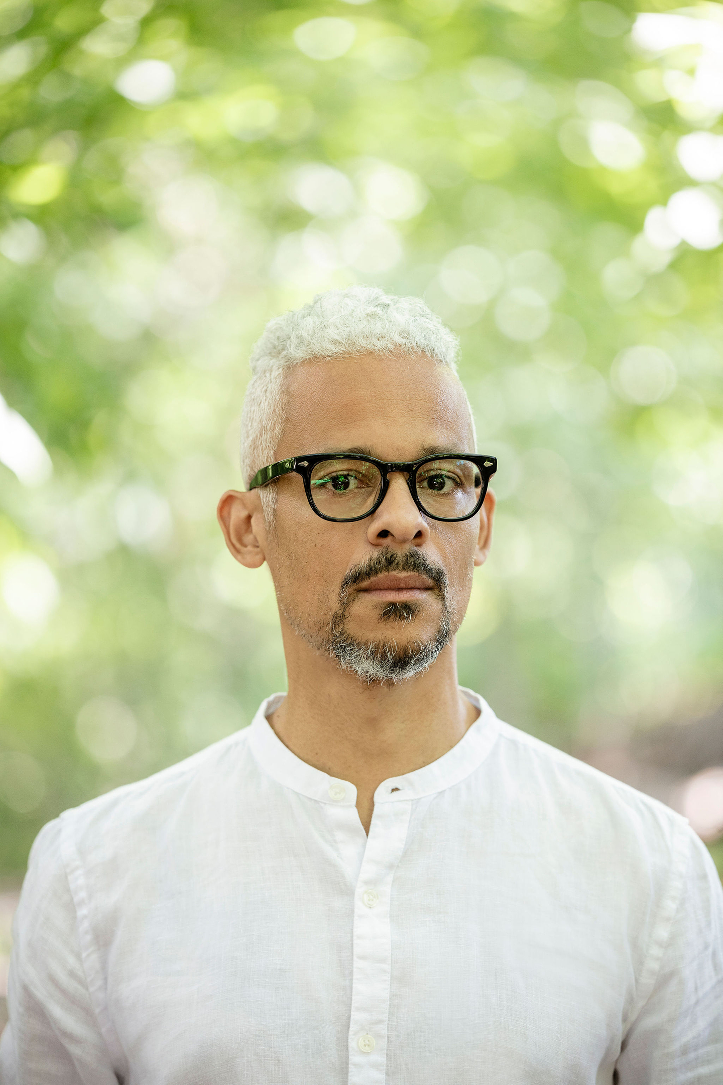 Kaie Kellough holds a soft expression while gazing into the camera. He has short silver hair, and wears black framed glasses and a white mandarin collar shirt with the top button undone. He stands outdoors with blurred green foliage and dappled light in the background.