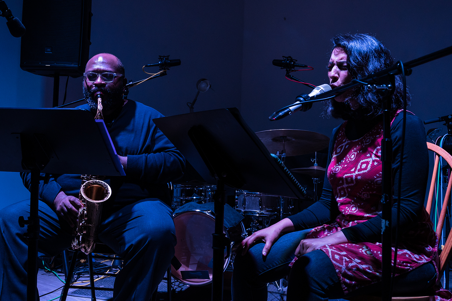 Two musicians are seated in a dark space surrounded by instruments and recording equipment. One is playing the saxophone and the other is singing into a microphone. An atmospheric blue light partially illuminates the face of the singer, whose eyes are closed and eyebrows are furrowed, giving the impression she is completely absorbed in the performance.