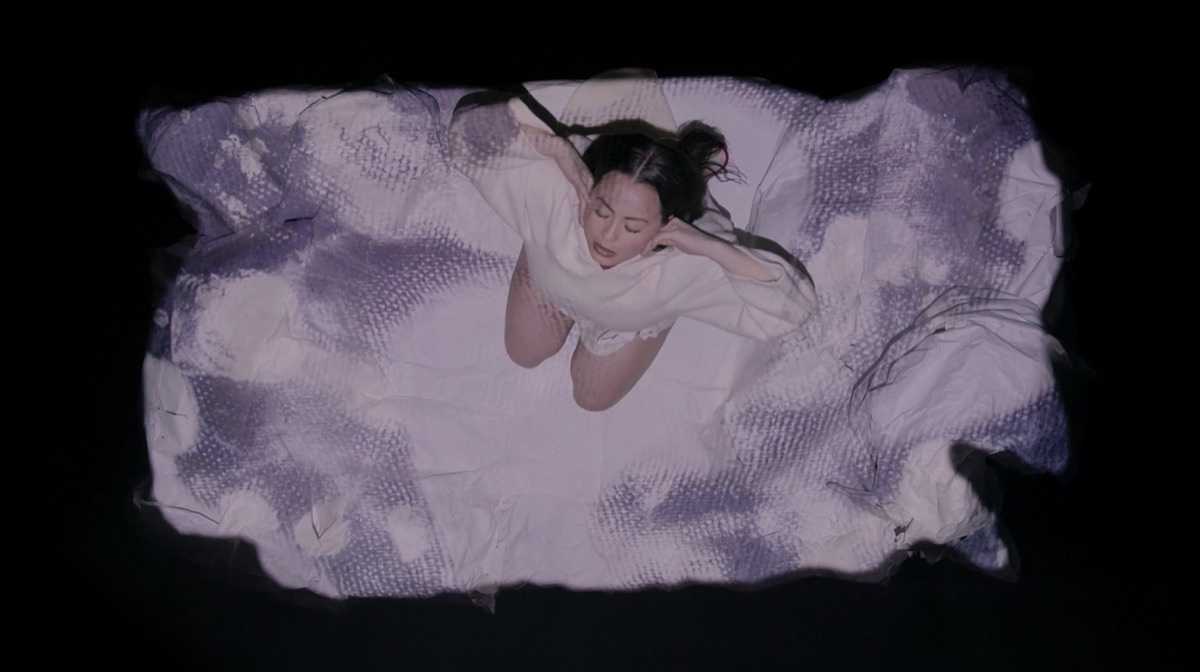 A photograph of Tiffany Ayalik performing Itqaqhaijuq / Tries to Remember at Western Front. The photograph is taken from a bird’s-eye point of view above Ayalik as she kneels on the sculptural set for her performance, a white rectangular shape resembling snowy ground or floating ice. She wears a white wool amaut – traditional Inuit woman’s coat. Ayalik tilts her head up with eyes closed, her elbows outstretched, and her hands resting on her cheeks. 