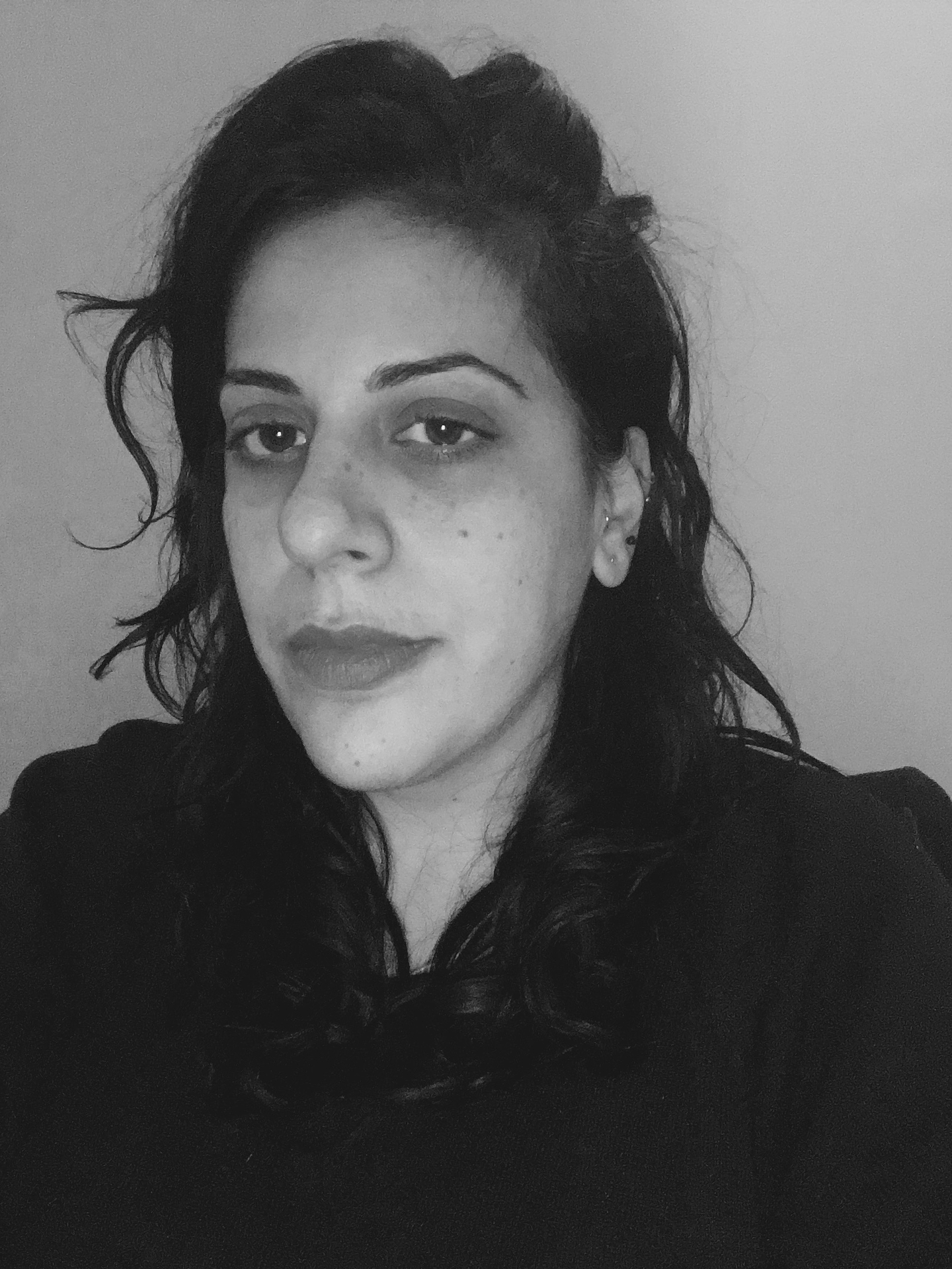 Black-and-white portrait of Suzy Halajian, a woman with dark curly hair worn loose over her shoulders and gathered at the top, looking directly at the camera with a neutral expression. She wears a dark, high-necked top and small stud earrings, photographed against a plain light-gray background.
