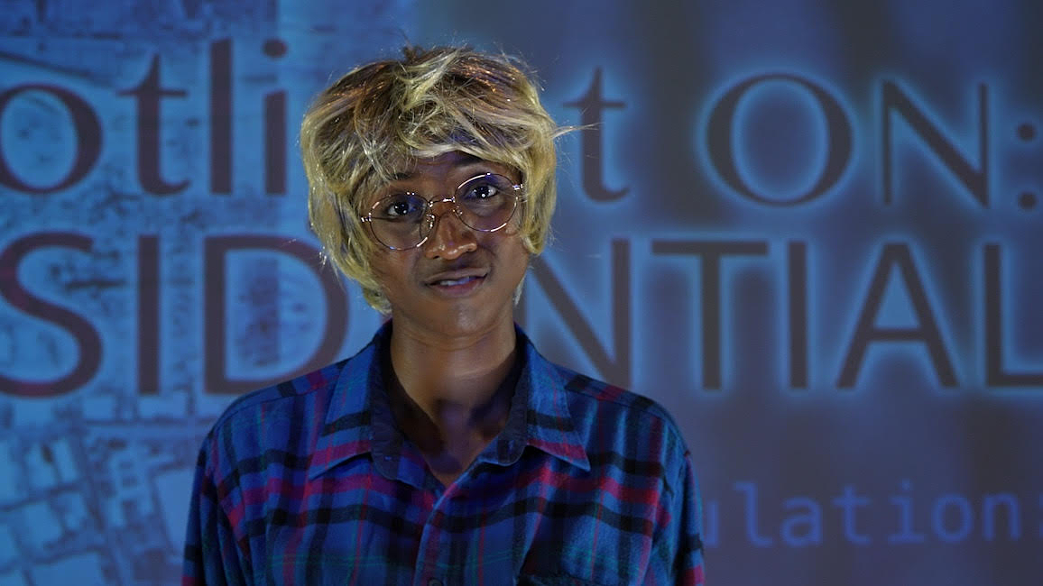 Lex Brown, wearing a blue plaid shirt, glasses, and cropped blonde wig, stands in front of a blue background of layered digital images including a satellite photograph of buildings near a river.