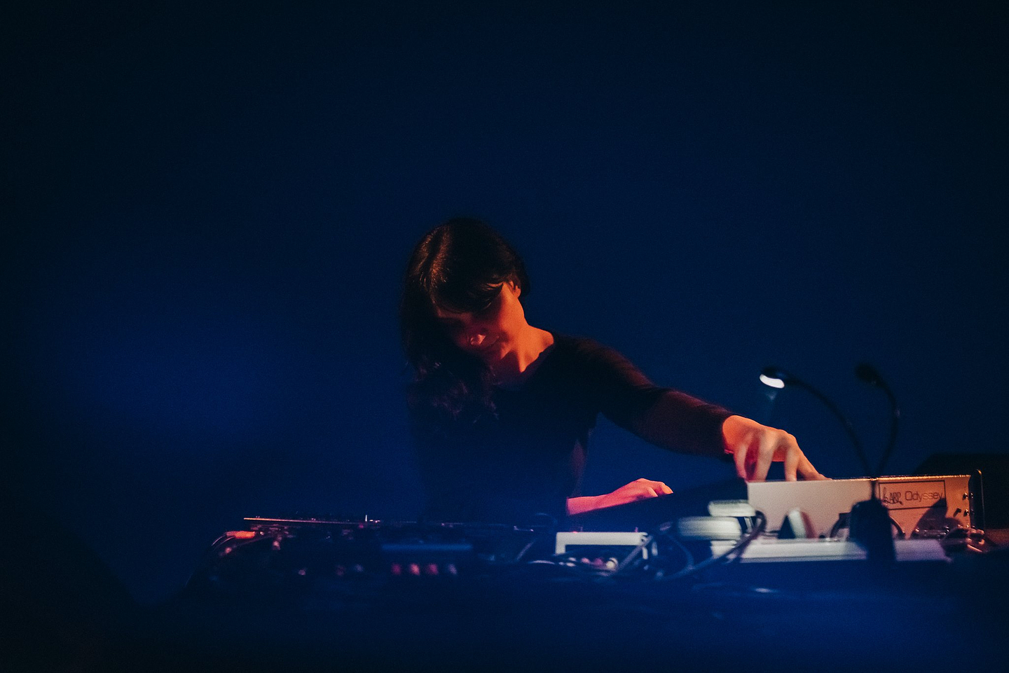 A woman wearing a dark coloured shirt is standing in front of an electronic music setup. Her left hand is adjusting a knob on a mixing station. She is faintly lit by a blue light.