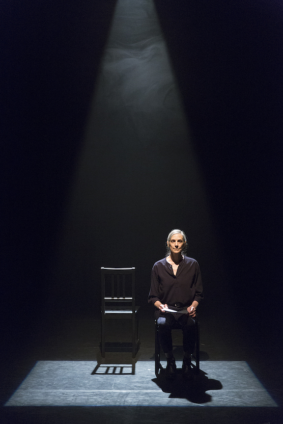 A colour photograph of Peggy Baker seated on stage looking directly towards the camera. She is dramatically spotlit from above, and in her hands she holds a sheet of paper. To her left is an empty black chair.