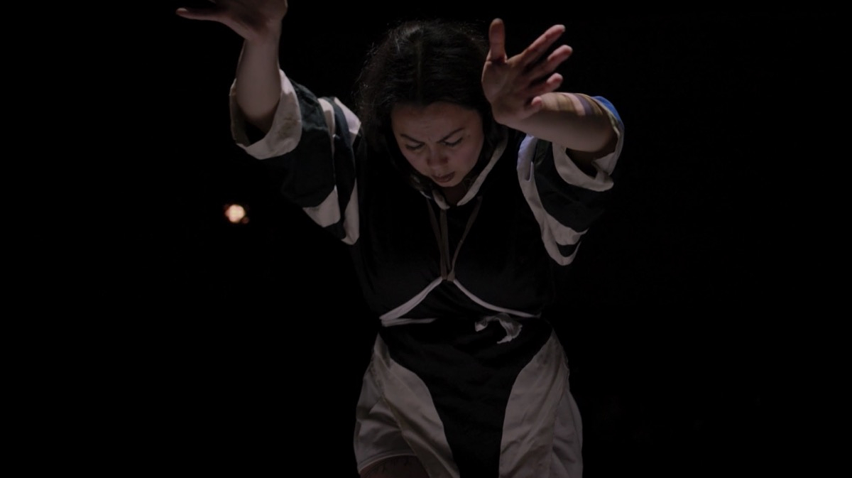 A photograph of Tiffany Ayalik performing Itqaqhaijuq / Tries to Remember at Western Front. Ayalik is wearing an amaut – traditional Inuit woman’s coat – with black-and-white patterns, and a white rope-like belt tied across her chest and around her waist. Her arms are stretched out toward the camera and her face is tilted down.