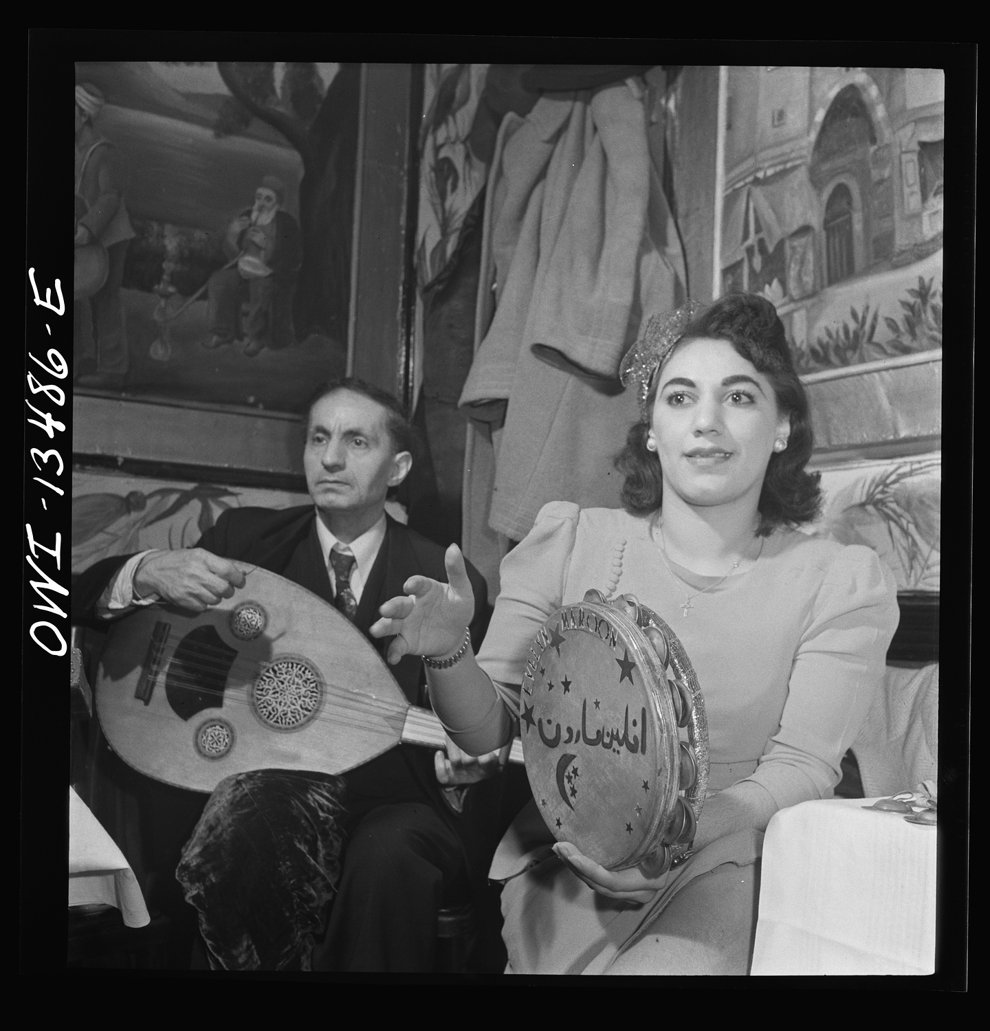 A black-and-white photo of a man and a woman seated in the corner of a club. The man wears a suit and plays the lute. The woman wears a fascinator and a long-sleeved dress, and plays the tambourine.