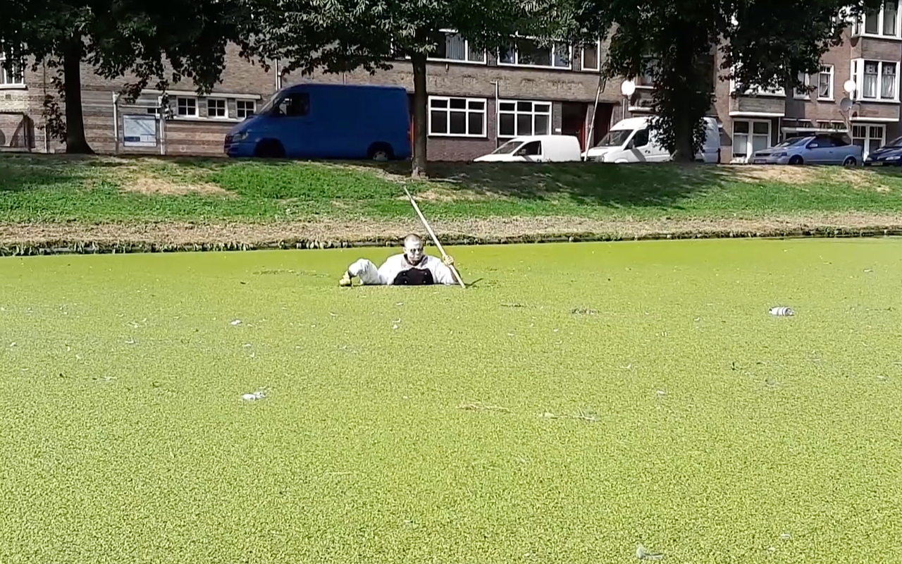 Geo Wyeth stands chest-deep in a canal covered in duckweed, wearing a baggy white shirt and holding a long pole.
