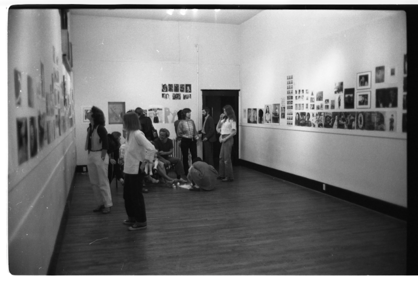 A black and white photograph of the interior of Western Front’s gallery, in which an exhibition of many photographic portraits of different sizes, tightly crowded together on the walls, is on display. There are visitors in the gallery, some looking at the work on the walls and some seated on the floor.