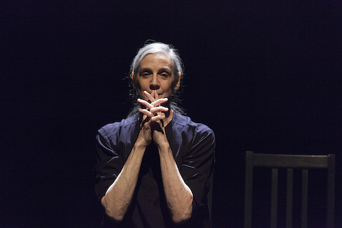 A colour photograph of Peggy Baker performing. She looks directly towards the camera and holds her hands together with fingers interlaced to her lips. To her right is an empty black chair. 