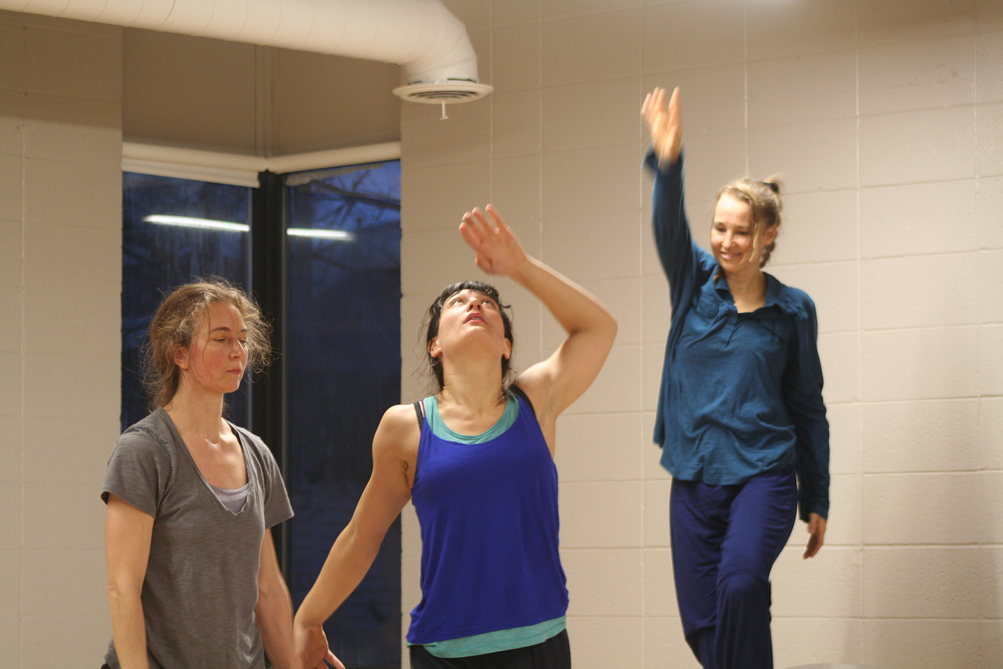 Three dancers stand next to each other in a well lit room. The dancer on the left stands with her arms by her side and her eyes closed as if in deep meditation. The dancer in the middle holds her left hand above her head and looks upwards with a sense of momentum, while the dancer on the right is elevated and raises her right arm upwards creating the impression that she is lifting up the dancer in the middle through this action.