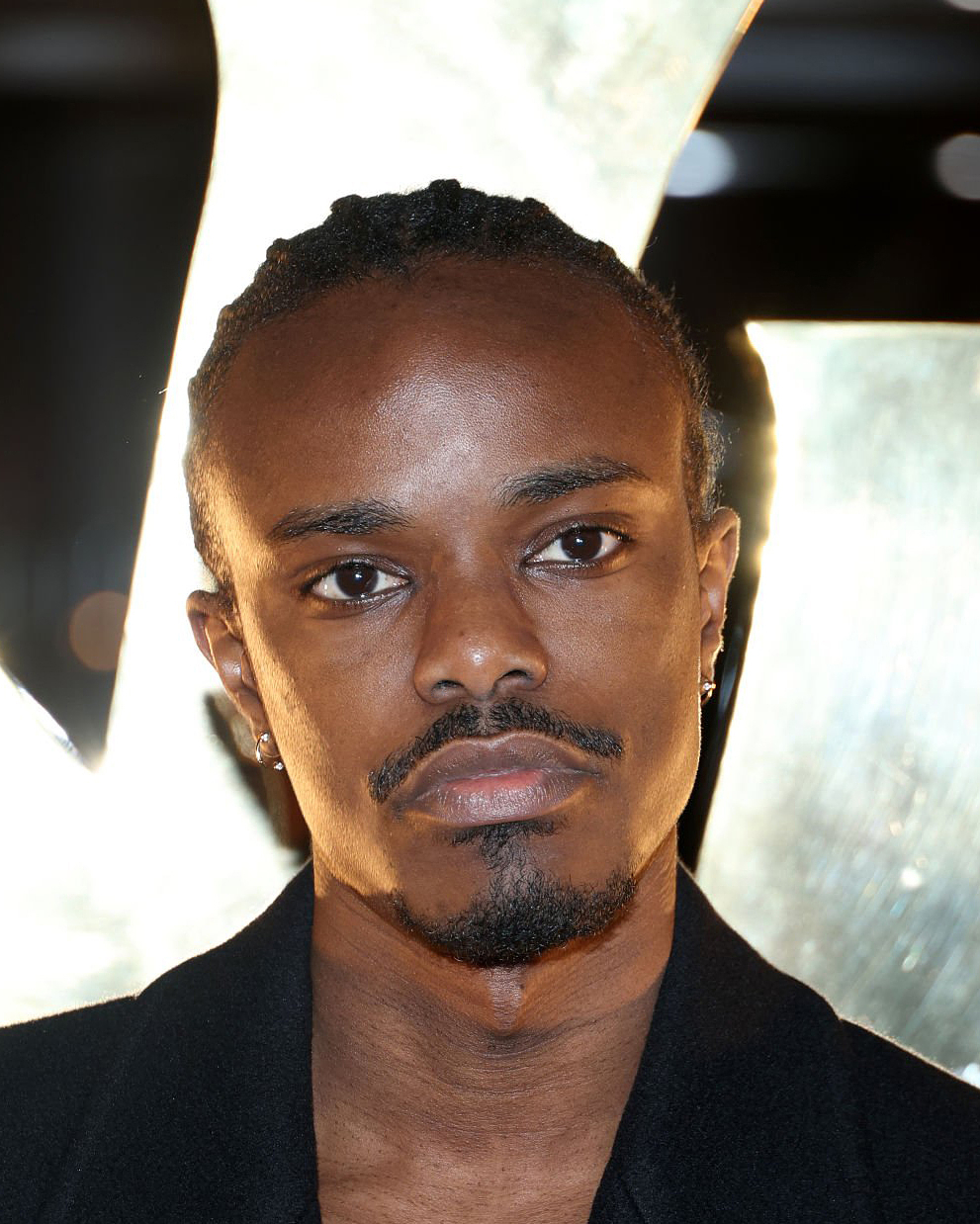 A person with short, tightly braided hair is photographed from the shoulders up, standing in front of a large, bright, out-of-focus light shape that creates a glowing halo effect behind their head; the person is wearing a dark collared garment.