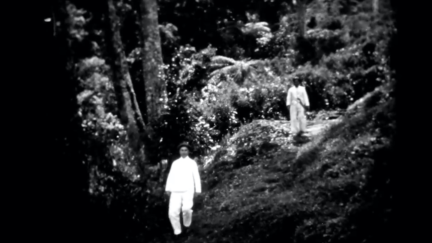 A black-and-white film still depicting two people dressed in white walking downhill in a lush forest.

