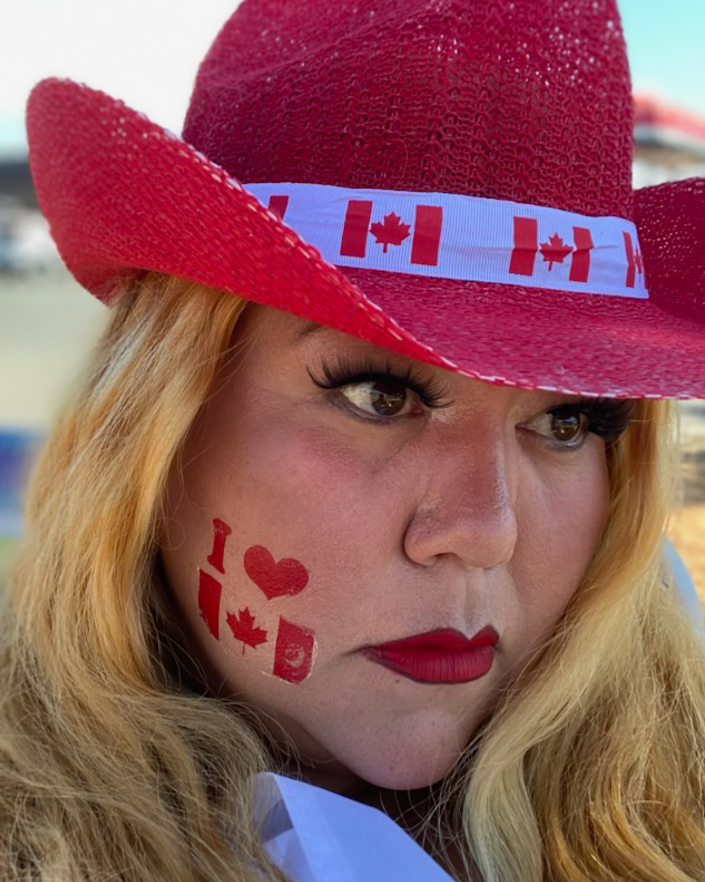 Close-up portrait of Skeena Reece, with long blonde hair, false lashes and red lipstick, gazing sideways toward the camera with a serious expression. She wears a red woven cowboy hat with a Canadian flag ribbon band, and her cheek has a temporary tattoo reading "I ♥" over a Canadian flag. Behind her, out of focus, are a blue sky and a parking lot.