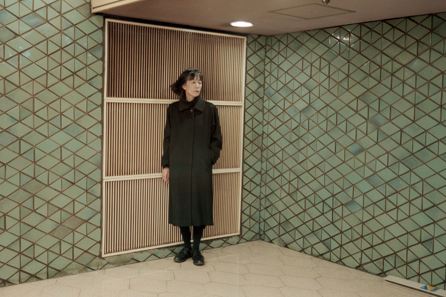 Phew stands in front of a cream-coloured ventilation grill installed in a wall featuring a pattern of green triangle and diamond tiles. She wears all black and looks over her left shoulder. 