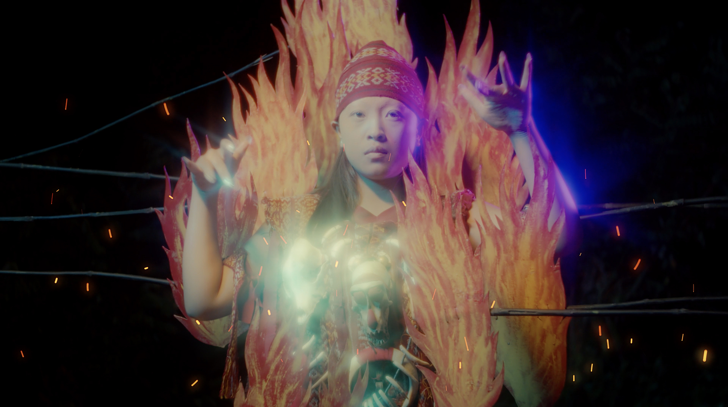 Surrounded by animated flames, a young femme presenting person dressed in traditional red kabasaran attire raises their arms while gazing towards the camera. Red, yellow, and orange sparks glow against the image’s black background.