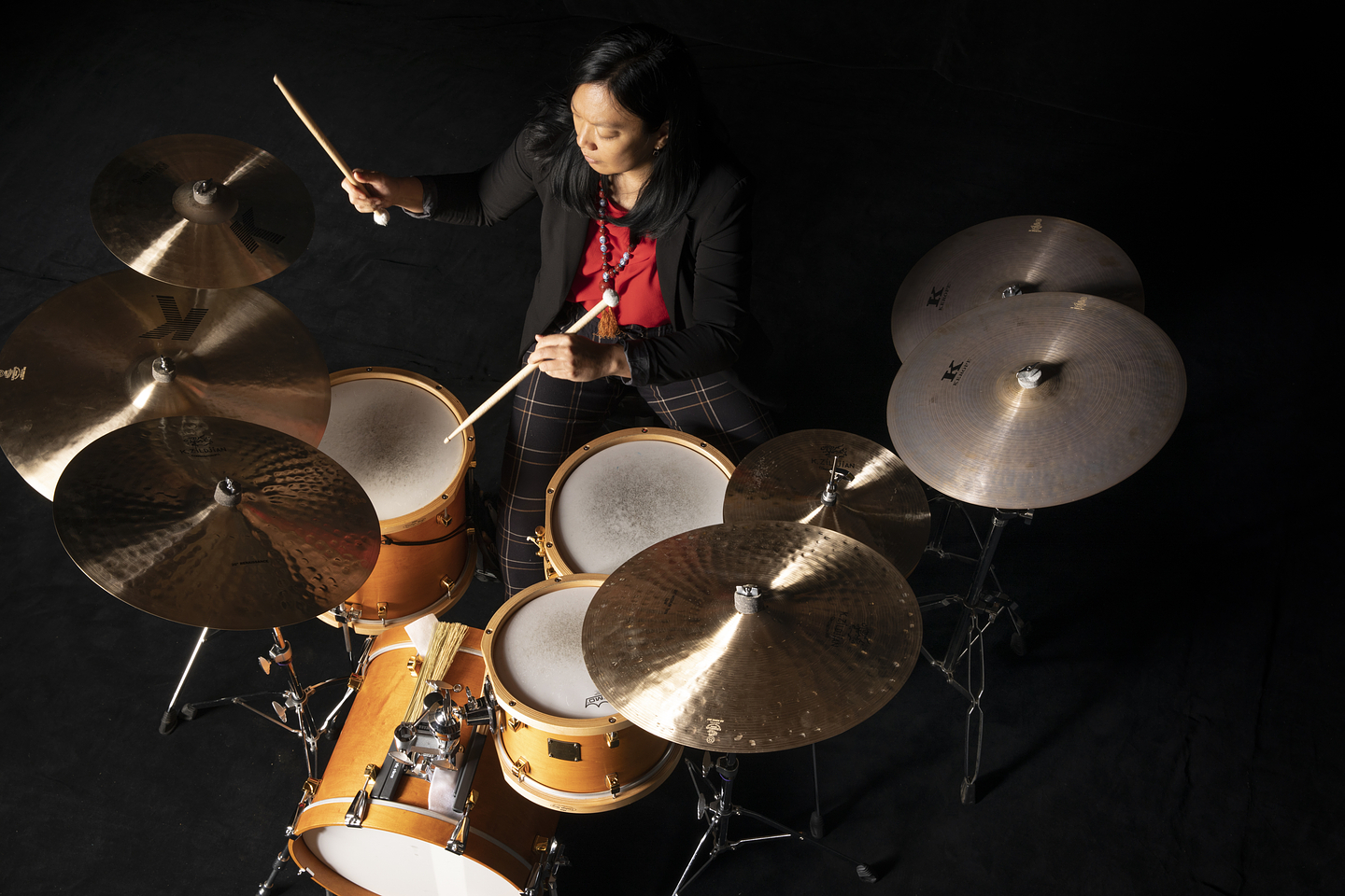 Susie Ibarra plays a drum set from an overhead perspective. She is dressed in a black blazer, red top, and plaid pants, holding drumsticks mid-motion. The drum kit includes multiple cymbals, toms, and a snare, all arranged on a dark stage background.