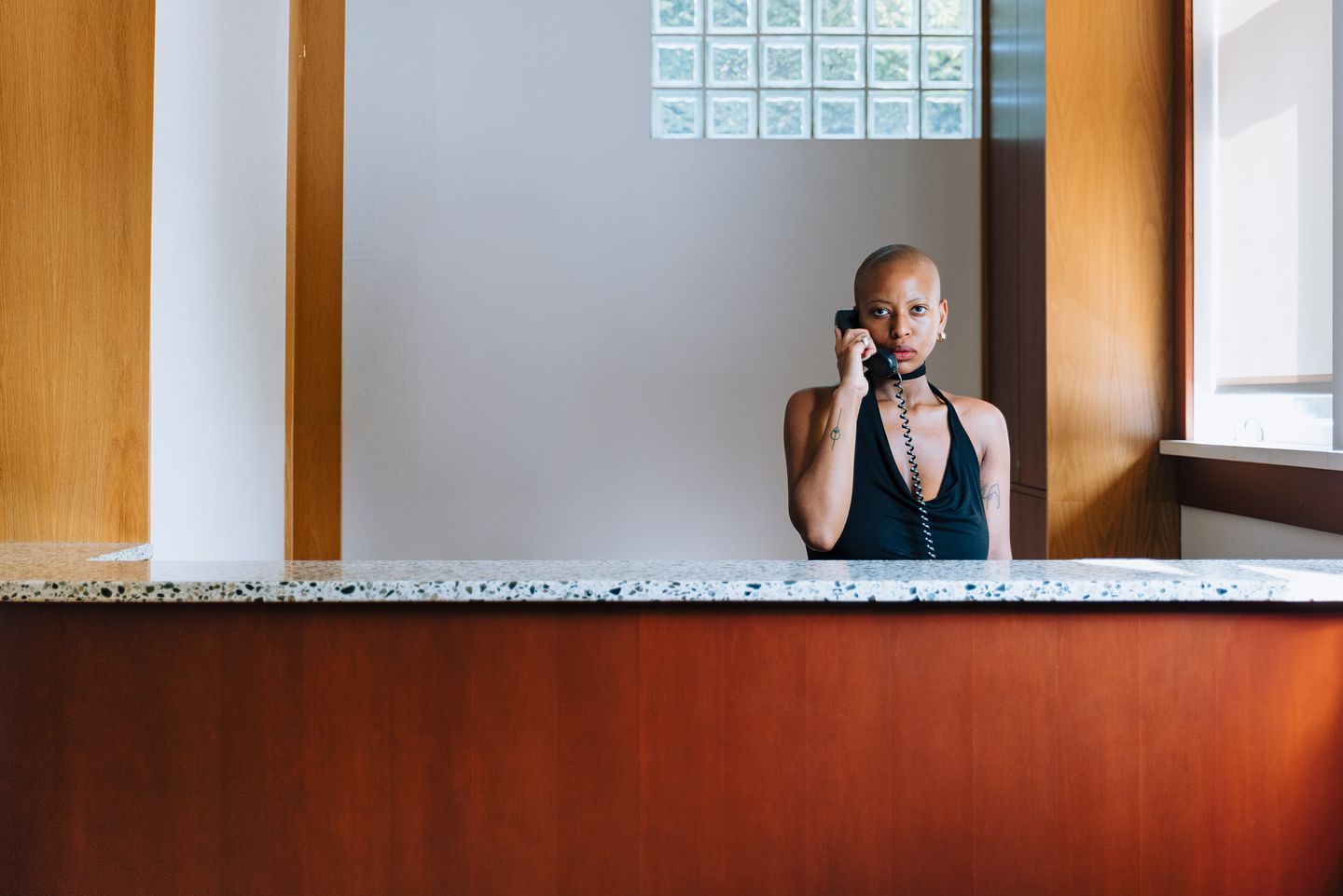 Chipo Chipaziwa stands behind a reception desk holding a black telephone to her ear. She wears a black halter-top with a deep neckline and meets the camera's gaze directly. The desk has a speckled marble top, and the background features light wooden panels and a glass block window, allowing soft daylight to filter into the space. 