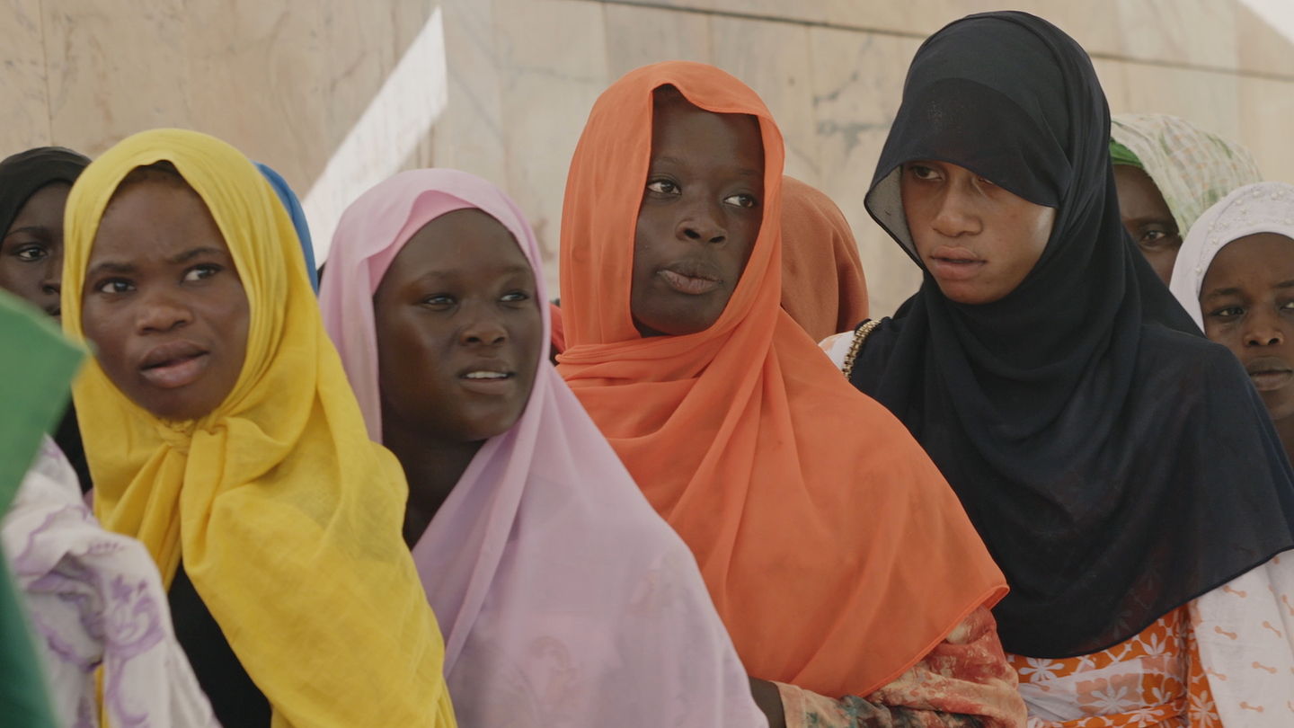 A group of young women stand close together in sunlight against a pale stone wall, wearing headscarves in yellow, pink, orange, black and white. Most look off to the left with attentive, serious expressions, as if listening to or watching something outside the frame.
