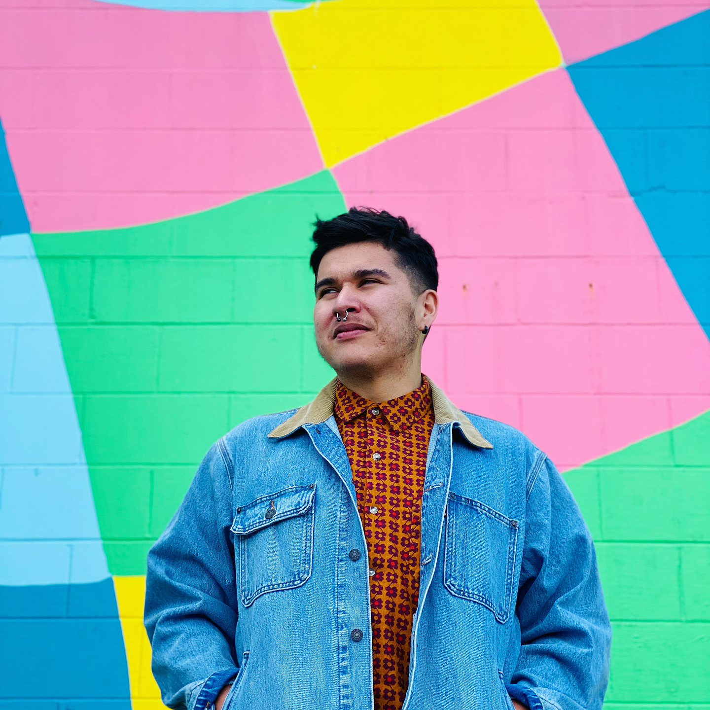 A portrait of Billy-Ray Belcourt standing in front of a wall painted in a colourful geometric pattern.