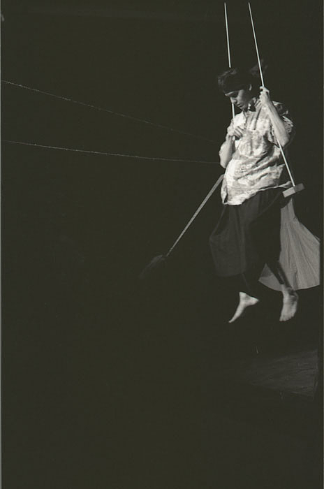 Black and white portrait oriented image of an Indigenous man on a swing, lit by a spotlight. He wears a dark coloured headband that holds back his dark hair, a light coloured patterned short sleeve button up shirt, a dark coloured skirt, and no shoes. He sits on a swing made of a simple plank suspended by two white ropes that extend out of the frame at the top of the image. He holds the left side rope with his left hand, his right arm is wrapped around the right side rope and pinches his shirt at the chest. He gazes slightly downward, his eyes concealed in shadow. Behind him a white dress hangs from a line. The rest of the stage is dark.
