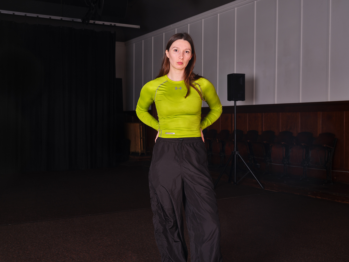 Nina Davies stands in theatrical space. She looks directly at the camera with her hair down and hands placed behind her back. She wears black tracksuit pants and a bright lime coloured sports top.  