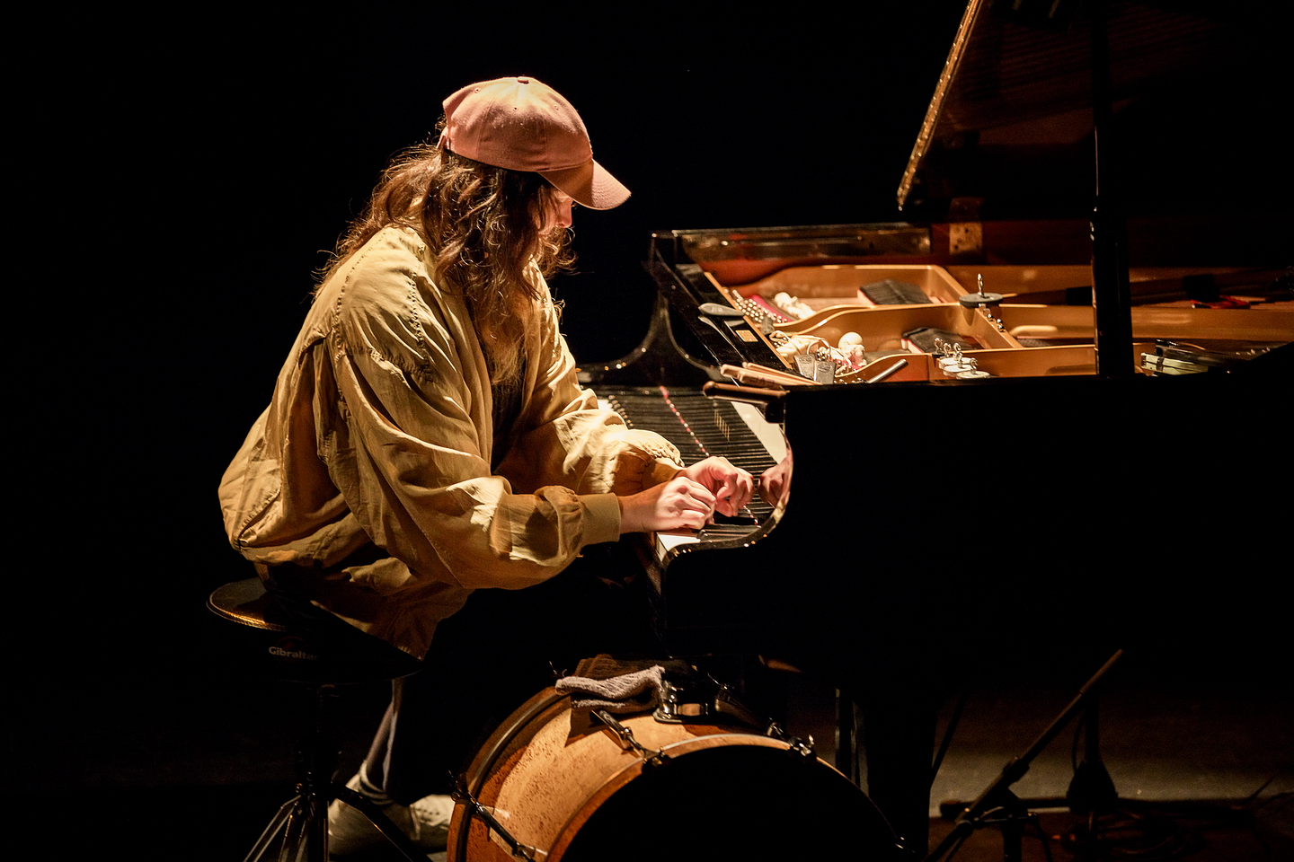 Eve Risser wears a loose beige jacket and a pink baseball cap while playing a grand piano on a dimly lit stage. She is seated on a drum stool, with a large bass drum positioned below the keyboard. The piano's lid is open, revealing various objects placed inside the instrument, suggesting a prepared piano setup. The scene is intimate and atmospheric, with warm lighting highlighting the performer’s focused posture.