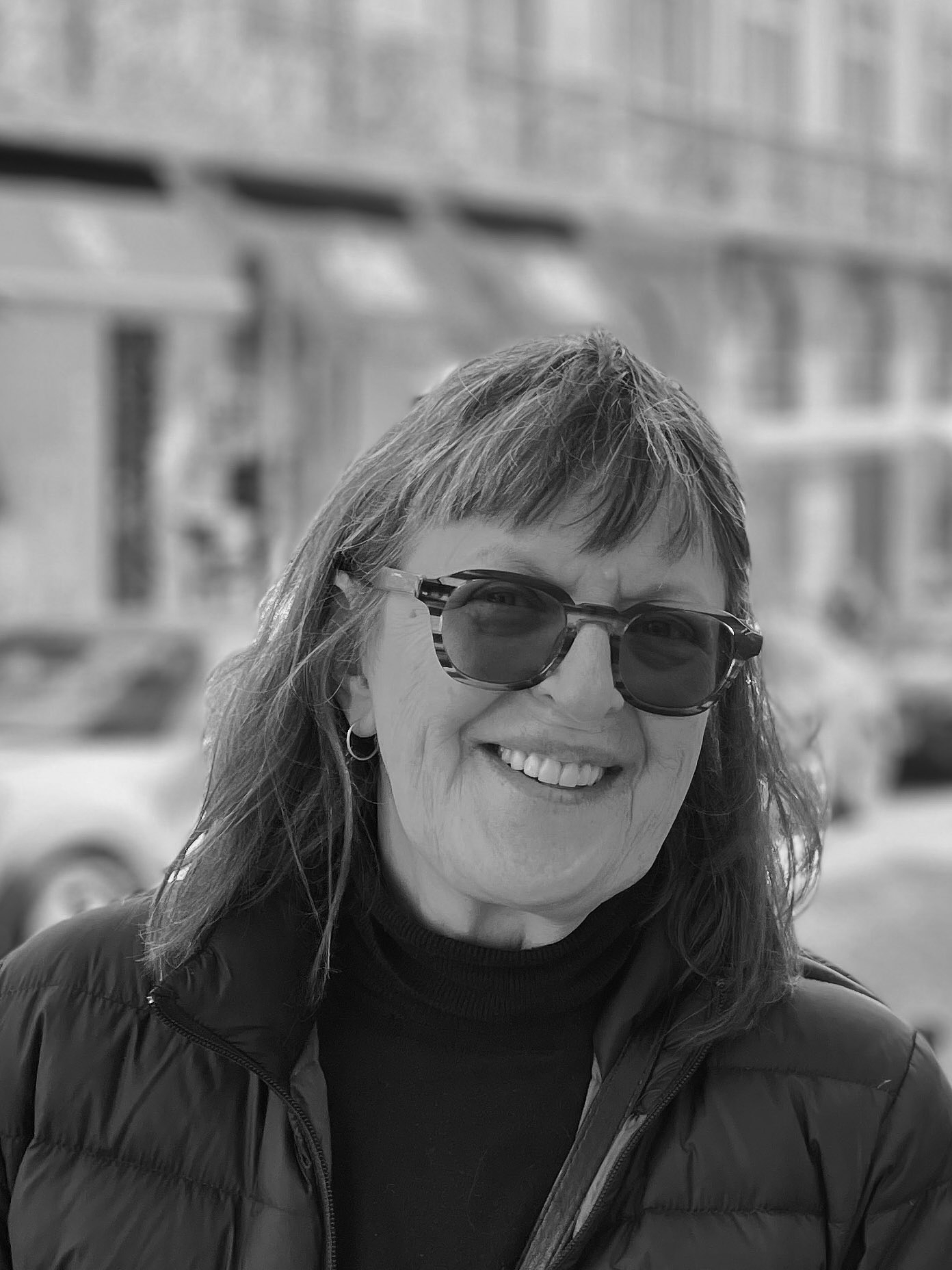 Erin Moure smiles at the camera. She wears transition lenses, hoop earrings and a puffer jacket. The street behind her is blurred