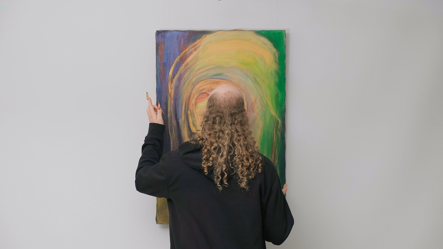 Archie Barry wears a black hoodie and stands facing a colorful painting with swirls of yellow, green, and purple. They hold up one hand with fingers slightly raised, appearing to engage with the artwork.