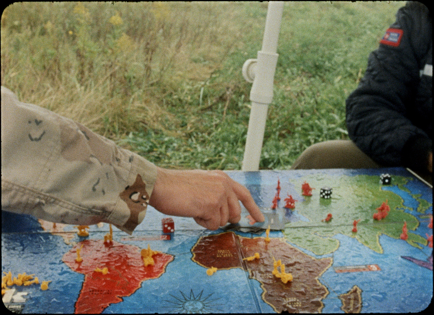 Close-up of a person in a camouflage jacket pointing at a world map game board with various coloured pieces, while another person in a black jacket sits across. They're seated outdoors, with a grassy field seen behind them.
