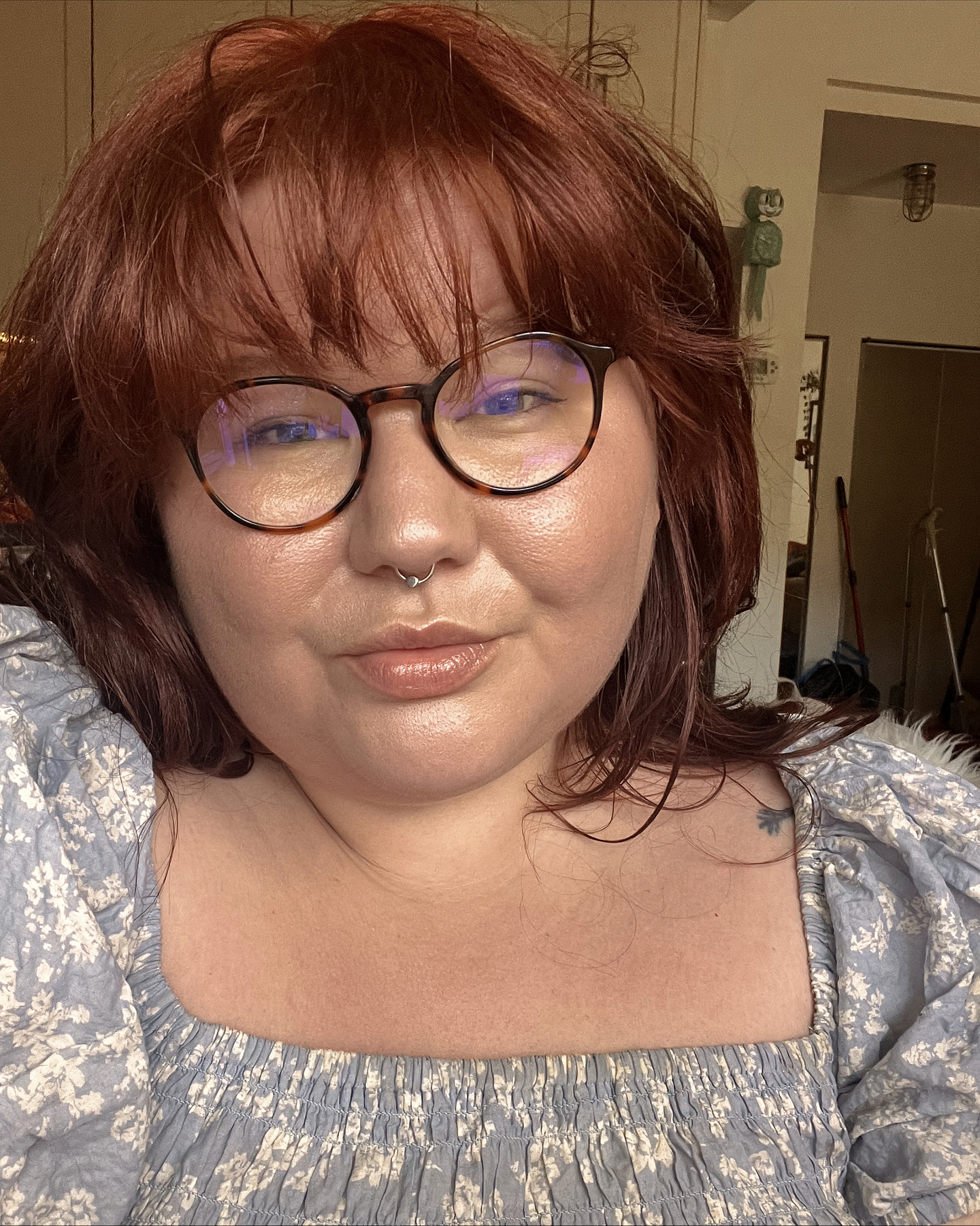 Samantha Marie Nock wears round tortoise shell glasses, a silver septum piercing, and a blue and white patterned ruched peasant top. She is seated indoors, and a green Kit-Cat Klock can be seen hanging on the wall in the background.