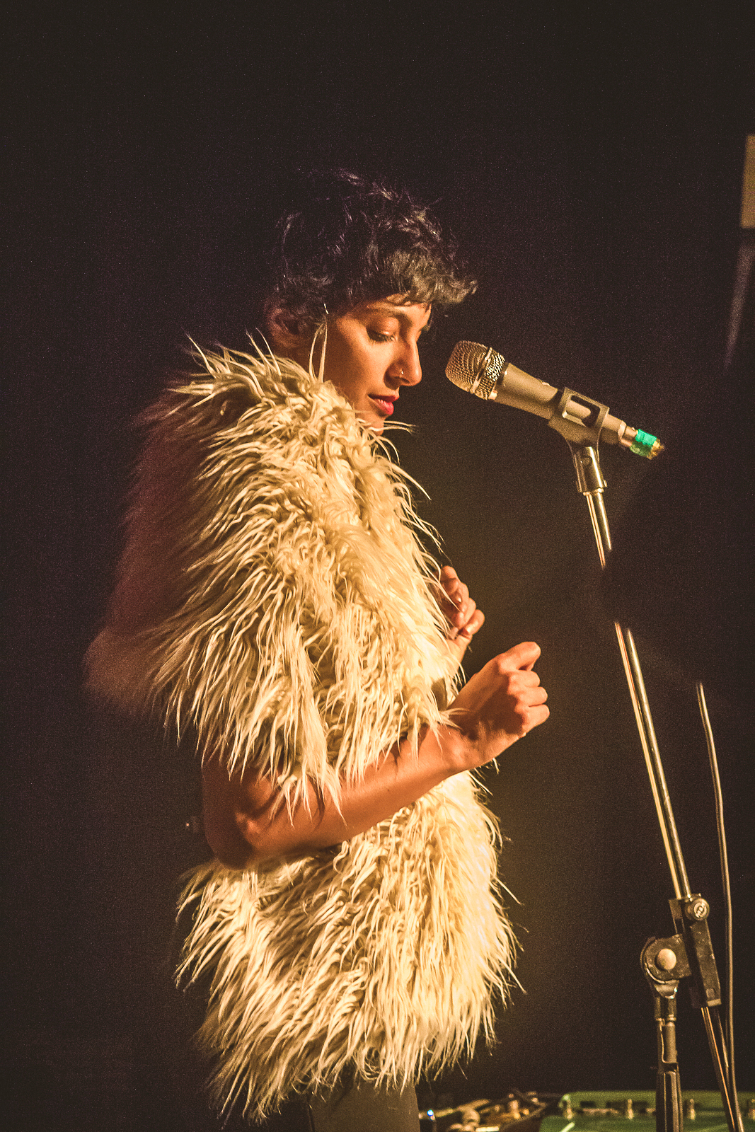 Samita Sinha is seen from the side standing next to a microphone on a stand. She is poised in preparation for singing. Around her shoulders she wears a shaggy white shawl.