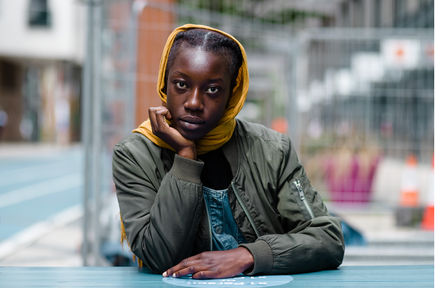 Samra Mayanja leans against an outdoor table with her chin resting on her right palm. She wears an olive green bomber jacket and a yellow scarf that is wrapped around her neck and tucked over her head. 