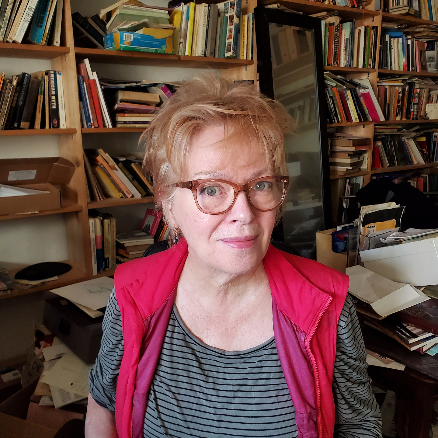 Dorothy Trujillo Lusk sits in a cozy, book-filled room. The wall behind her is lined with shelves that are packed with a variety of books, papers, and objects. She has short blonde hair and wears glasses and a pink zip-up vest over a grey shirt with thin black stripes.