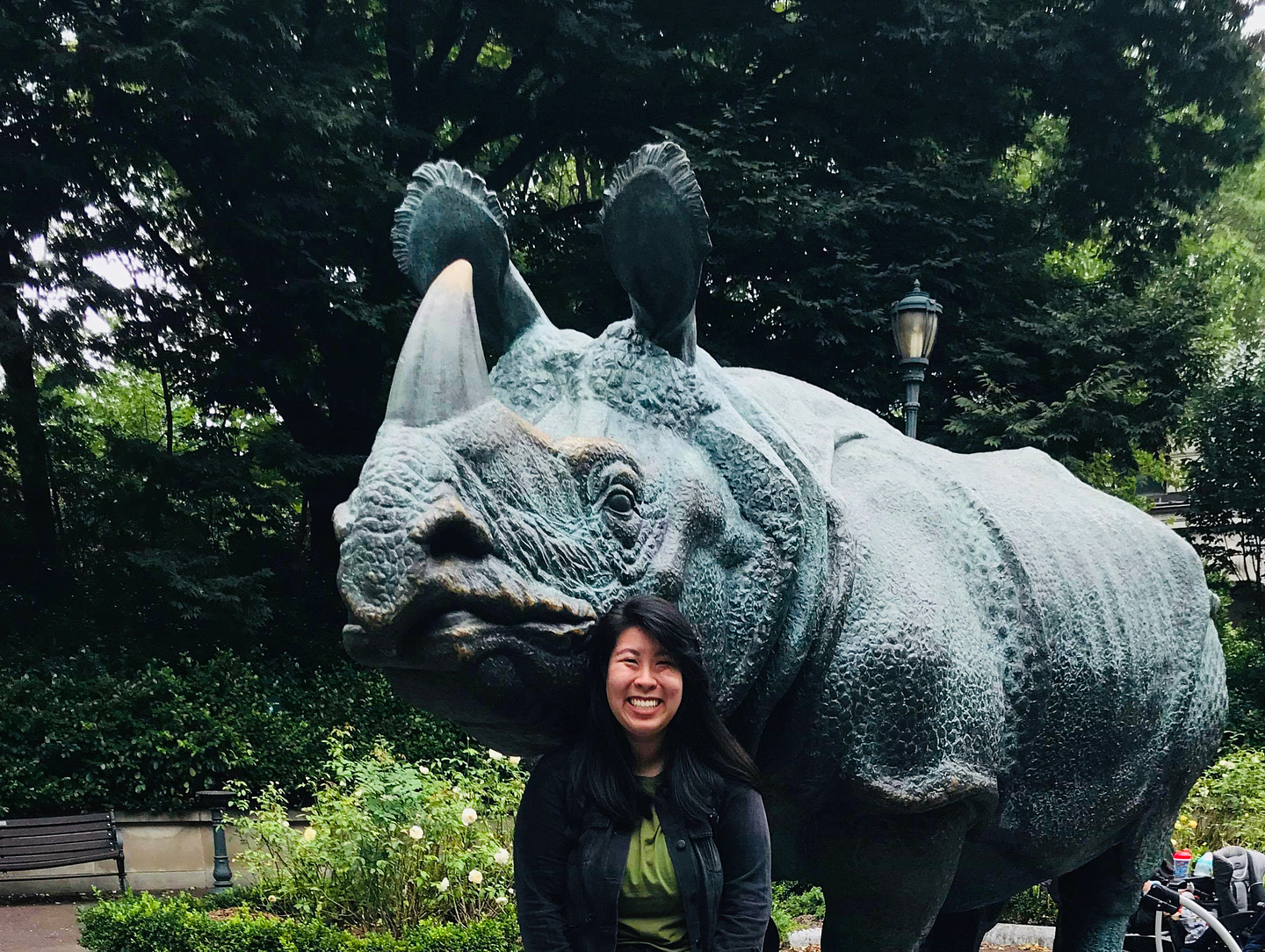 Smiling, Tina Do stands in front of a huge patinated brass rhinoceros statue in a public park. 