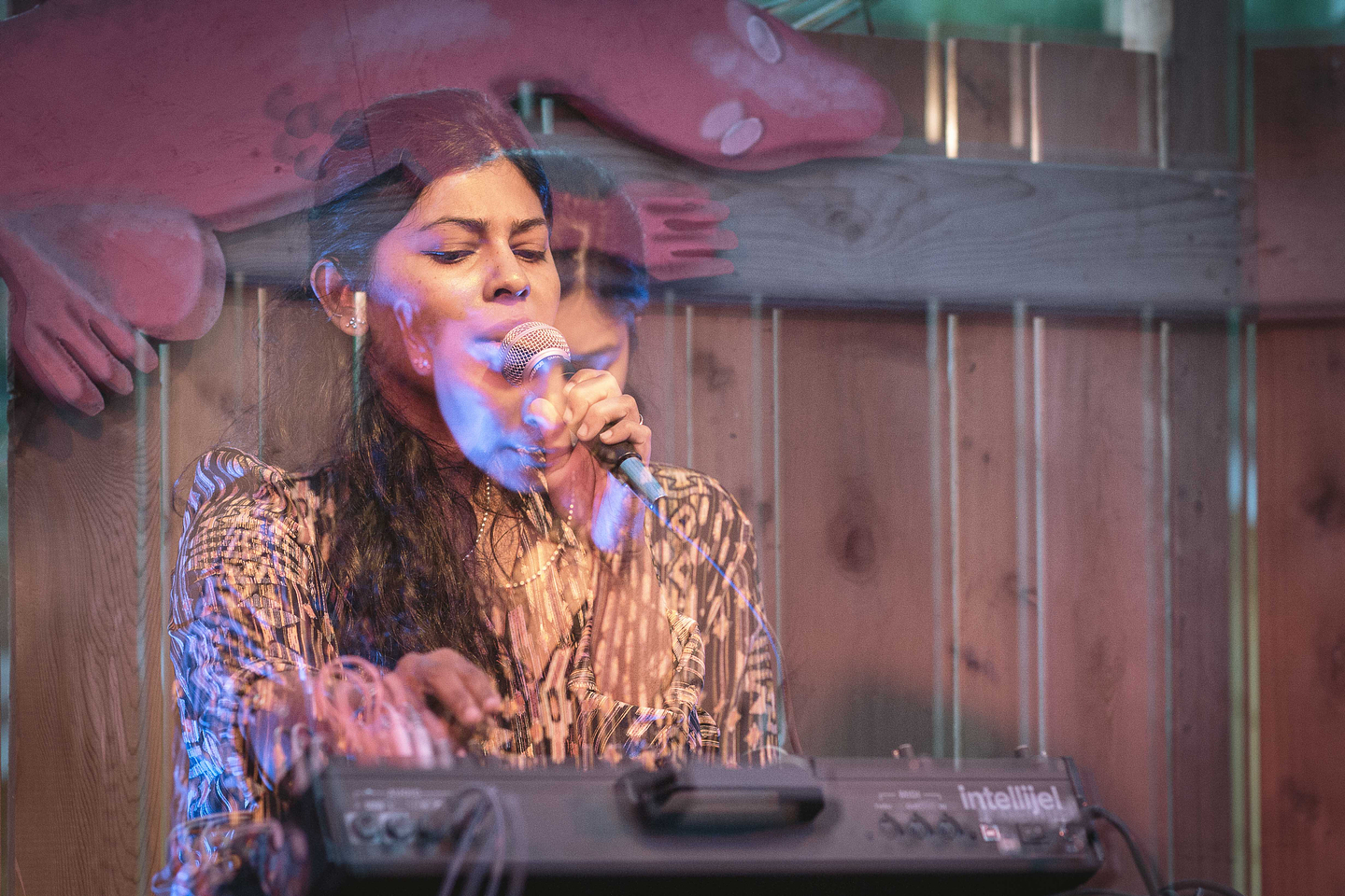 Piu sings into a microphone while playing an Intellijel synthesizer. The image has a double-exposure effect, creating a ghostly overlay of Piu in different positions. She wears a patterned shirt and has long dark hair. The background features a wooden fence and a large, abstract red figure, adding a surreal and vibrant atmosphere to the scene.