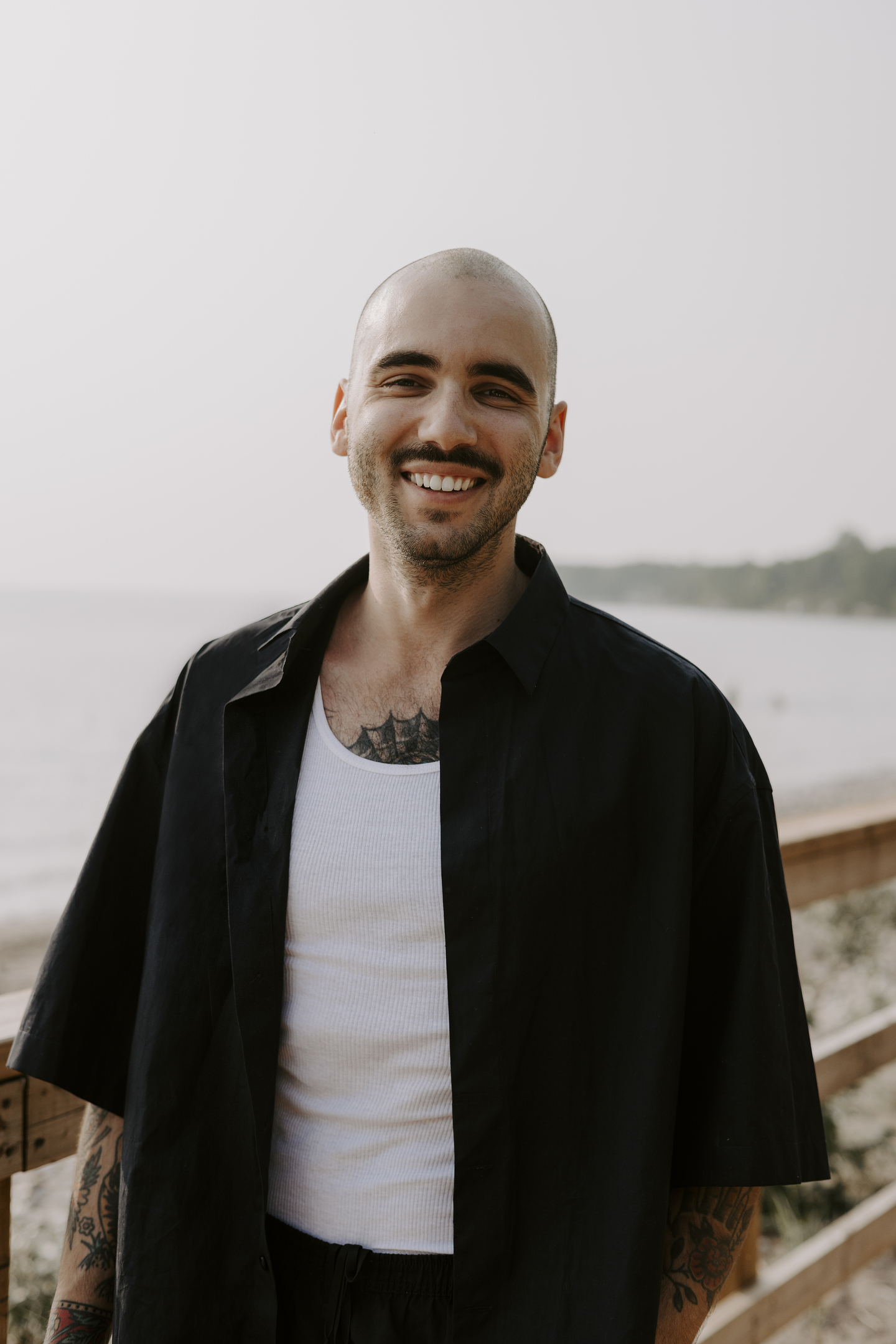 Cody Caetano smiles widely at the camera. He is posed on a pier with a body of water behind him, and wears a short-sleeve button-up shirt over a white tank top.