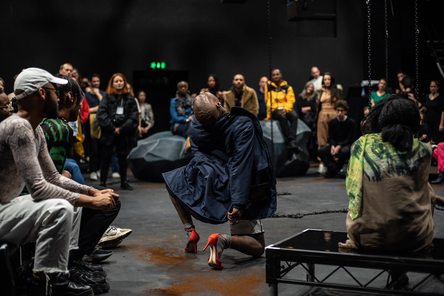 A colour photograph of keyon gaskin performing. keyon is kneeling in the middle of a black box theatre, with their left leg up, right knee on the ground, and their shaved head thrown back. They are wearing a navy trench coat and red high heel shoes. Surrounding them is an audience seated on low rise staging.