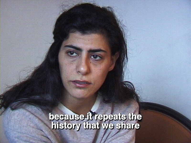 Souha Bechara is seated and looking to the left. She has long dark hair that rests on her shoulders. She is wearing a grey sweater with a white collar and clip on microphone. The subtitles, because it repeats the history that we share, appear in white text near the bottom.