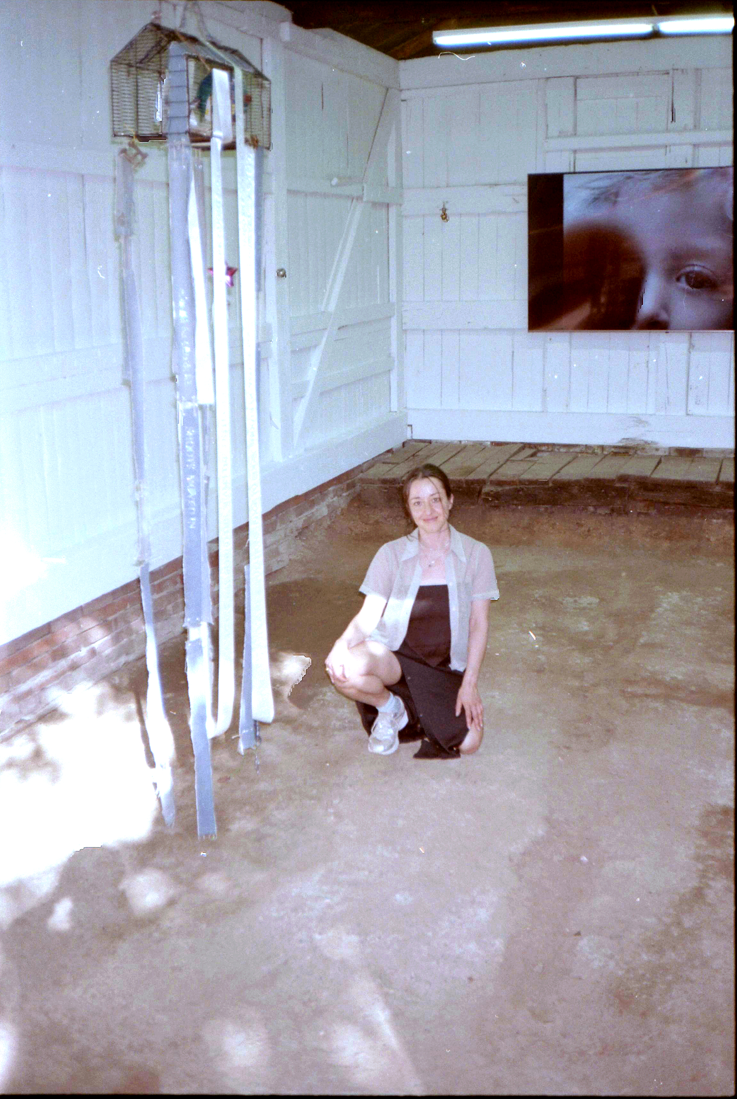 Smiling, Tiziana La Melia kneels on the floor of a rustic, white-painted room with wooden walls. A monitor showing an image child's face is mounted on the wall behind her. To her left hangs a sculpture consisting of a birdcage adorned with long strips of ribbon. 






