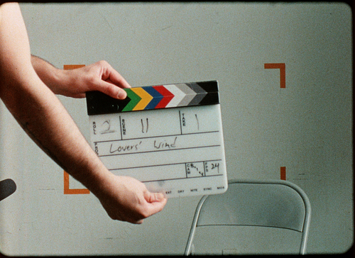 A clapperboard is held centre frame, featuring the words "Roll 2, Scene 11, Take 1, Lovers' Wind" in black texta.