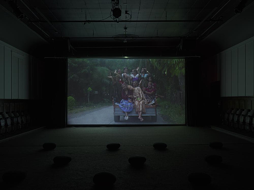 Ten people crowd the back of a pickup truck wearing traditional Filipino garments. Eight figures have their hands up in rejoice while artist Bhenji Ra rests her head on her mentor's shoulder. This is the focal point of the image.